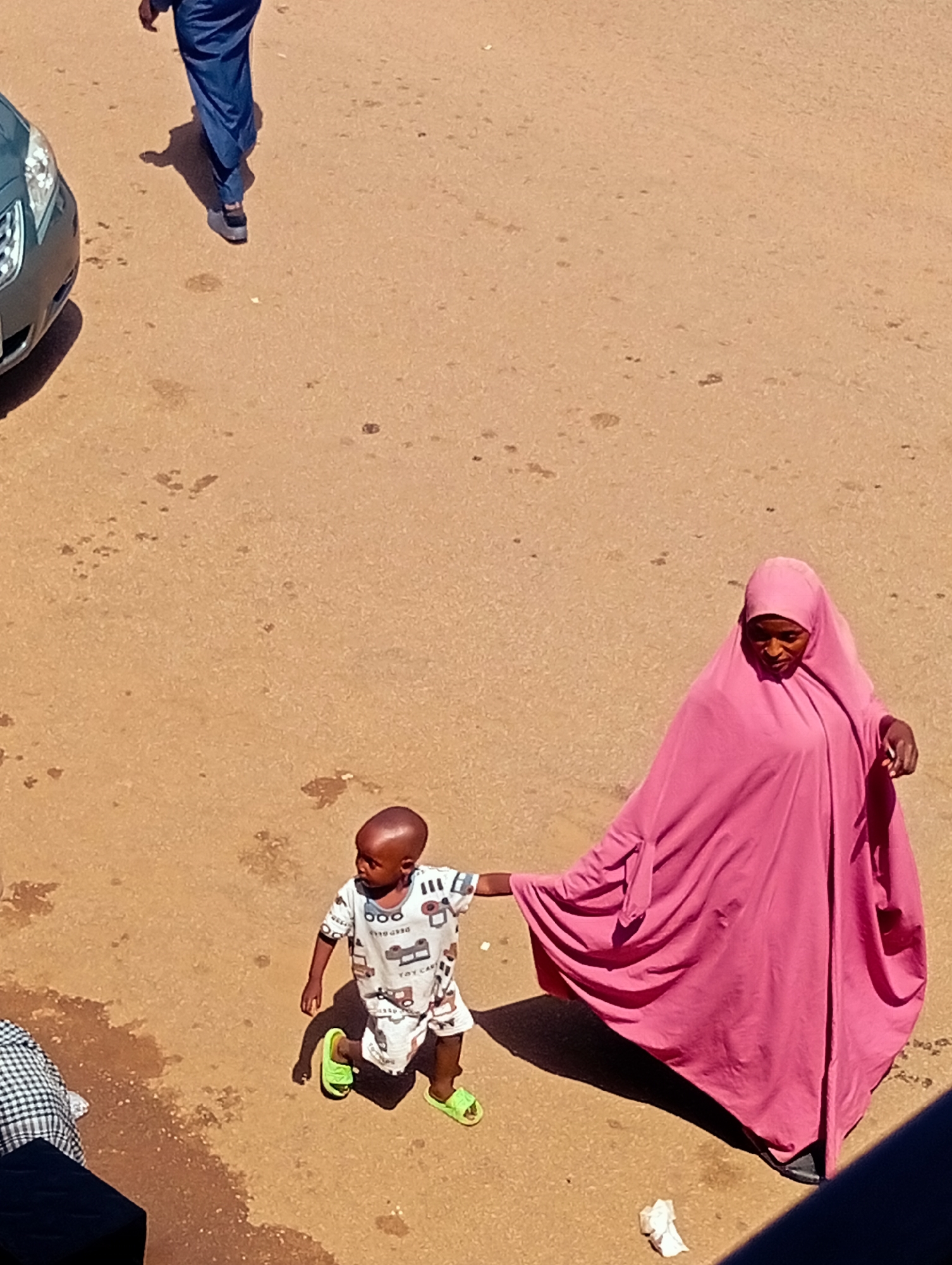 The image, taken from a high angle in Bukuru, Nigeria, captures a street scene with a child and an adult. A young child, wearing a white t-shirt with car prints and bright green sandals, stands on a dusty, light brown surface. The child is facing right, with a shaved head. To the right of the child, an adult, wearing a bright pink hijab and flowing robe, is partially visible. The adult's right hand is extended, touching or holding the child's left hand. The adult is looking downwards. In the background, on the left side of the image, the front of a dark gray car is visible. Further back, on the upper left, a person wearing dark blue trousers is walking away from the camera, casting a shadow on the ground. The ground appears to be a mixture of sand and dirt, with some scattered debris, including a crumpled white paper item near the adult.