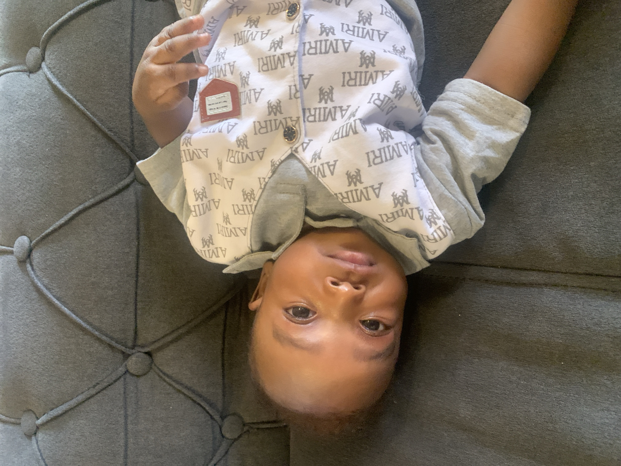 A baby is lying on a gray tufted couch. The baby is wearing a light gray and white outfit with the word "AMIRI" printed repeatedly, alongside a logo. The baby's head is oriented towards the bottom of the frame, while the feet are at the top. The baby's eyes are open, and the gaze is directed toward the camera. The background is the gray fabric of the couch. No other details relating to a location in Barakin Akawo, Nigeria are present in the image.