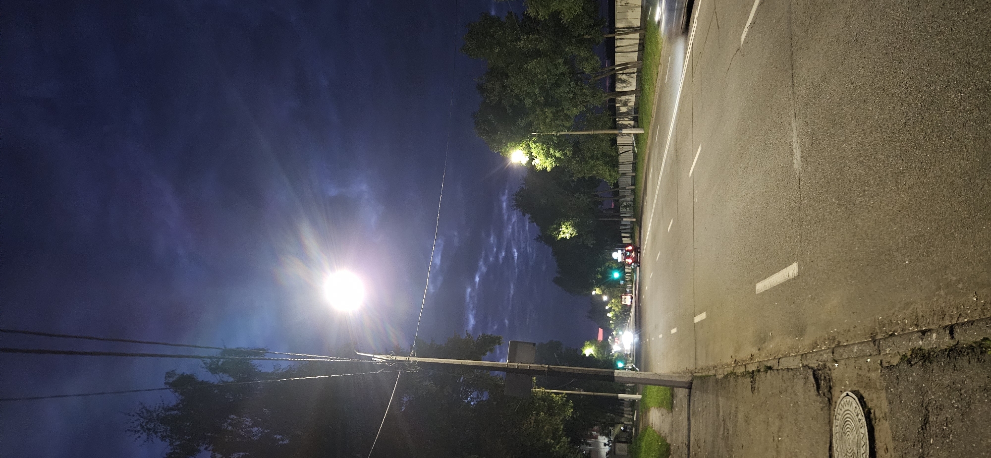 The image captures a street scene at dusk or night in Vitsebsk, Belarus. A bright, luminous streetlamp illuminates the foreground, casting a halo and creating lens flares that extend across the dark blue, cloudy sky. The street is paved and marked with white lane dividers. On the left side of the road, a sidewalk is visible, adjacent to a grassy verge and trees. A manhole cover is present on the sidewalk. Several cars are on the road, their headlights and taillights visible. Traffic lights, appearing green, are also visible in the distance. Trees line both sides of the street, and a white fence runs along the right side. The overall impression is of an urban environment transitioning from day to night, with artificial lighting playing a significant role in the scene.