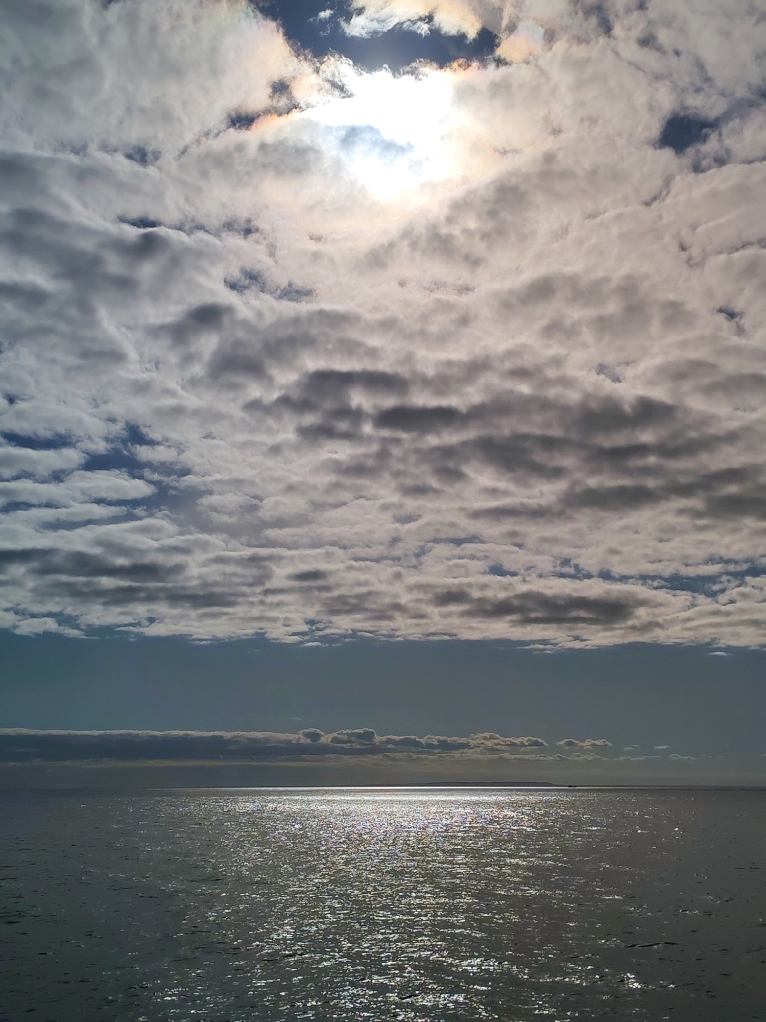 The image depicts a body of water, likely a lake or sea, under a sky filled with layered clouds. The sun is visible through the clouds, with a slight rainbow effect around it. The water reflects the sunlight, creating a shimmering effect. In the distance, a faint outline of land can be seen on the horizon. There are no people visible in the image. The exact location cannot be definitively determined as being in Ozernoe Ust'e, Russia, as the image contains no identifying markers specific to that location.