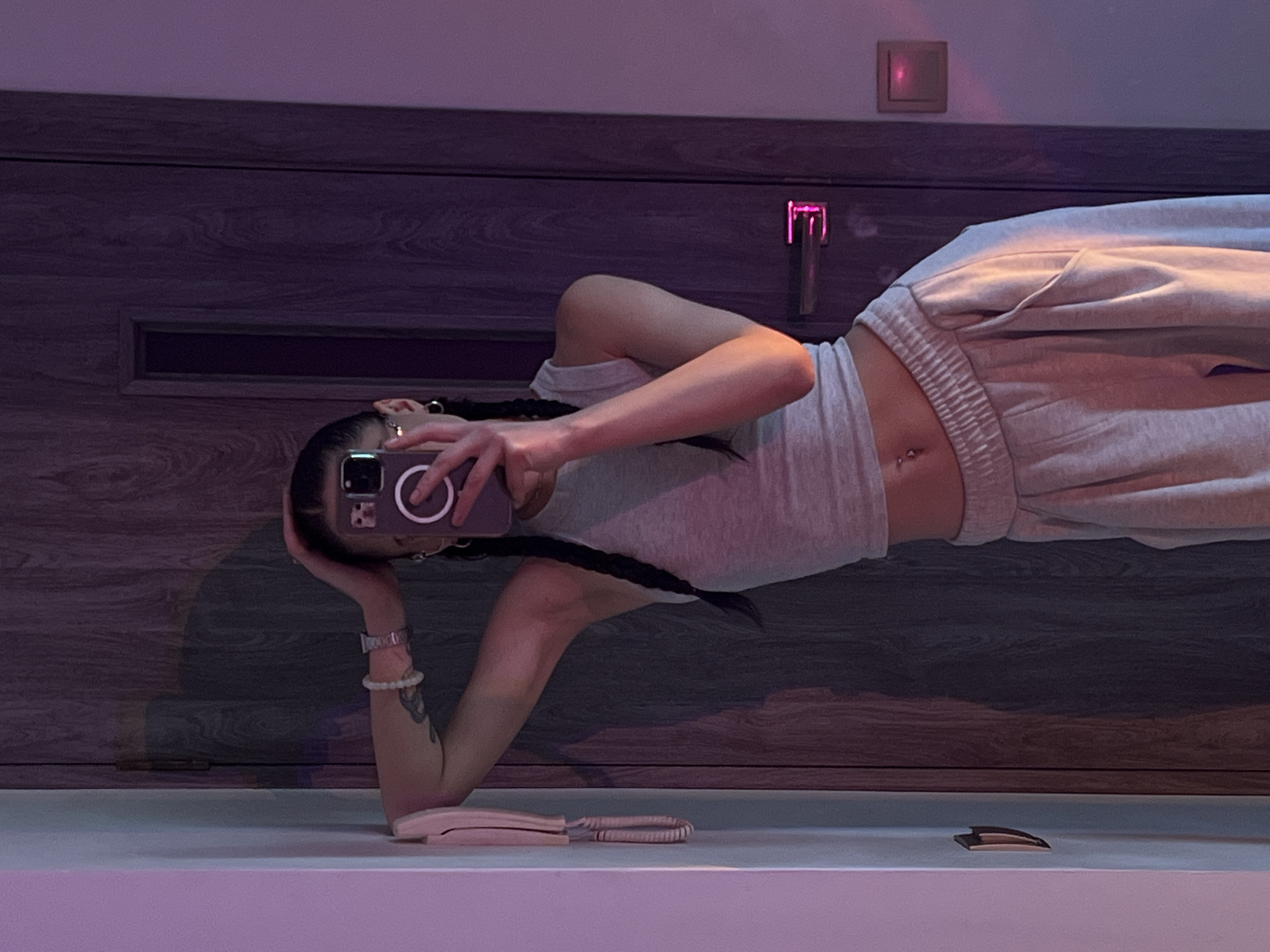 The image shows a person in a light grey tank top and light grey sweatpants standing in front of a dark wooden door. The person is holding a smartphone in their right hand, taking a selfie in a mirror. They have dark hair styled in two braids and are wearing a silver watch on their left wrist and small hoop earrings. The room has purple lighting and a light-colored wall with a mounted intercom device on the left. The wooden door has a modern silver handle with a pink light reflecting on it, and a small square light fixture is visible on the right side of the door. There is a slight shadow cast on the door from the person. The overall scene suggests an indoor setting, possibly a home or a hotel room, in Ternopil, Ukraine.