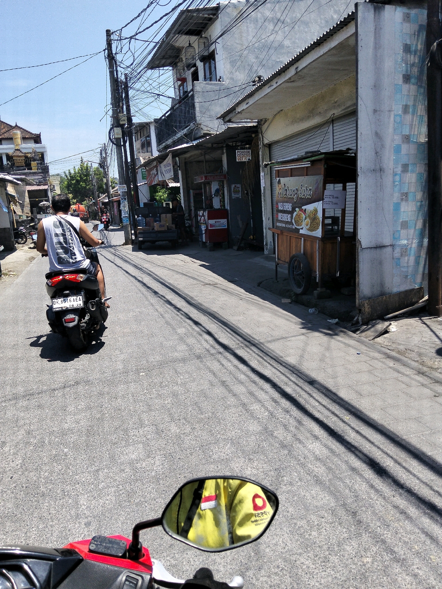 The image captures a street scene in Denpasar, Indonesia, on a sunny day. In the foreground, the handlebars and a red side panel of a motorcycle are visible, along with its rearview mirror reflecting a person wearing a bright yellow jacket with a logo. The rider of a black scooter, a man wearing a white sleeveless shirt and shorts, is the central subject, seen from behind as he travels down the asphalt road. The scooter has a license plate that reads "DK. 5516 AEA". The street is lined with buildings on both sides, featuring shops and food stalls. One stall, "Warung Sura", displays menus for "Nasi Goreng" and "Mie Goreng". Overhead, a complex network of electrical wires crisscrosses the sky, supported by utility poles. The pavement on the right side of the road is made of grey interlocking bricks. Shadows of the utility lines stretch across the road and pavement, indicating the direction of the sun. In the distance, other vehicles and buildings suggest a bustling urban environment.