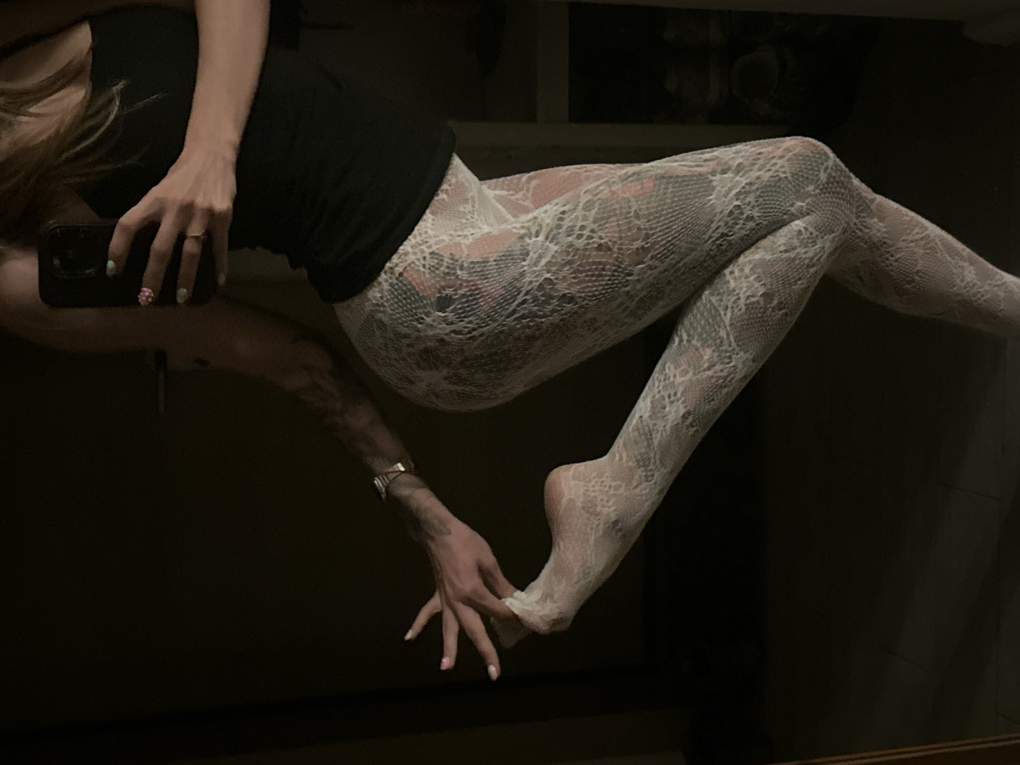 A person is standing in a dark room, possibly an apartment in Moscow, Russia. The person is wearing a black tank top and white lacy tights with a snake-like pattern. The person's left hand is touching their left foot, which is raised and also wearing a white lacy stocking. The person has tattoos on their arm. They are holding a smartphone in their right hand, which is reflected in a mirror. The lighting in the room is dim, with some light coming from a window or a light source not visible in the frame. The background is dark and out of focus, with some shelves visible on the right side.