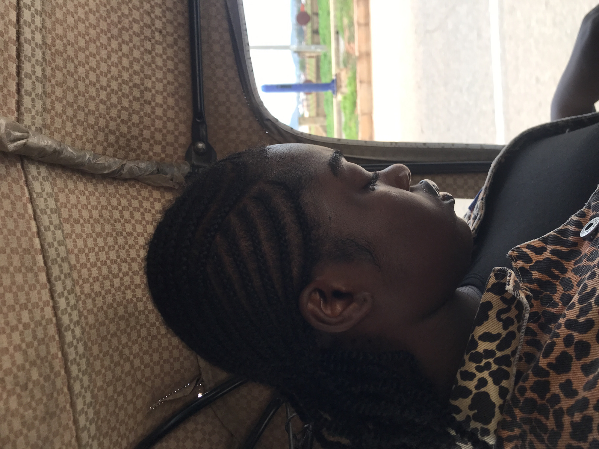 A dark-skinned female individual is captured in profile, seated within a vehicle in the city of New Stadium, Nigeria. Her hair is styled in cornrow braids, visible on the side of her head, leading towards the rear. Her gaze is directed downwards. She wears a dark-colored garment beneath a jacket featuring a distinct brown, black, and tan leopard print pattern. A single circular silver-toned button is visible on the jacket's front closure.
The vehicle's interior roof is lined with a fabric displaying a repeating grid pattern of alternating light and dark brown squares. Structural metal tubing supporting the roof is visible, with one section wrapped in a translucent, crinkled plastic material. Another dark metal bar runs horizontally across the upper frame.
Through the right-side opening of the vehicle, an external landscape is visible under a light grey sky. A light-toned paved surface occupies the lower portion of the exterior view, bordered by a distinct brown curb. Beyond the curb, green vegetation is present. A tall, slender blue pole or pillar stands vertically in the mid-distance. A low, light-colored fence or barrier extends horizontally behind the blue structure. Undulating landforms, indicative of hills, are visible on the distant horizon. Several indistinct reddish-brown circular shapes are also present in the far background.
