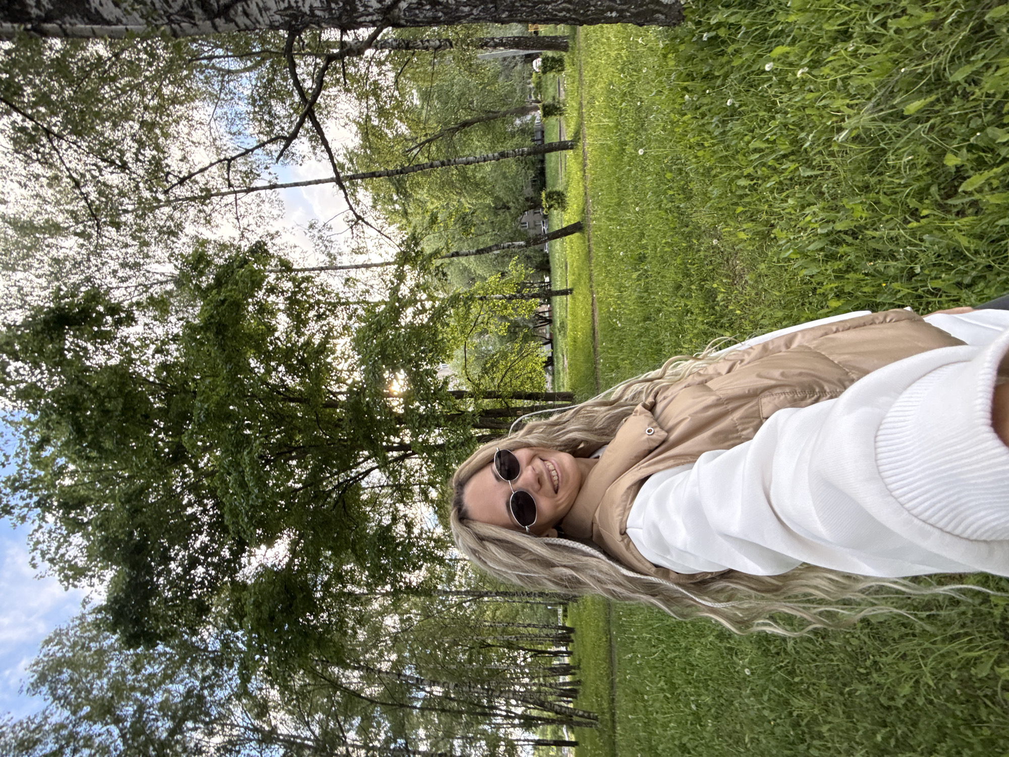A young woman with long blonde dreadlocks is lying on a grassy area in what appears to be a park. She's wearing sunglasses, a white hoodie, and a tan vest. The background includes trees, some of which are birch trees. The environment suggests a park or recreational area, possibly in Rusanovo, Russia, given the request. The photo is taken at an angle, likely a selfie. The lighting is bright, indicating daylight. There are no obvious interactions or actions, just a static pose.