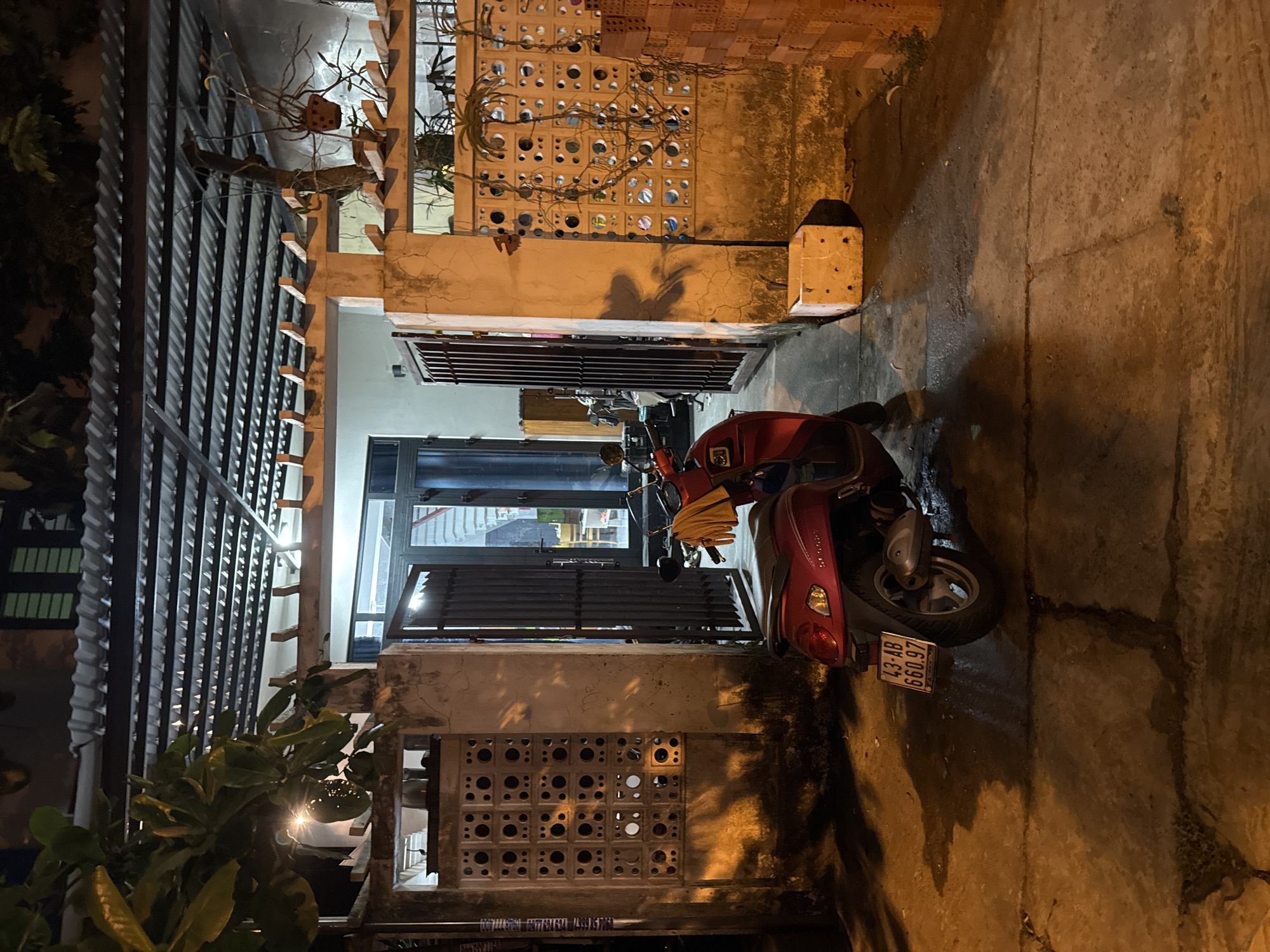 The image depicts a narrow alleyway in Đà Nẵng, Vietnam, at night. The alley is paved with concrete slabs, showing signs of wear and discoloration. A red scooter is parked on the right side of the alley, partially blocking the path. The license plate is visible, reading "43-AB 660.97".
Buildings flank both sides of the alley. On the left, a structure with a corrugated metal roof is visible, with plants spilling over the edge. A series of perforated concrete blocks forms a decorative wall. On the right, a similar concrete block wall is present, showing signs of weathering and staining. A doorway and window are visible further down the alley, with light emanating from within. Shadows are cast on the walls, suggesting a light source nearby.