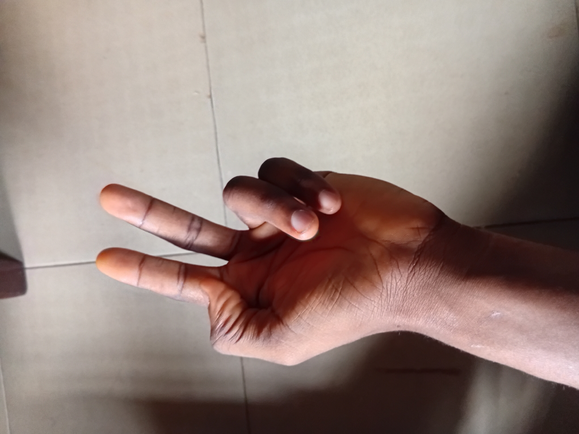 A person's right hand is shown from a slightly high angle, with the fingers extended upwards and the palm facing downwards. The hand is making a peace sign, with the index and middle fingers separated, and the thumb and ring finger closed. The skin tone is dark. The hand is positioned on a floor with light-colored tiles and dark grout lines. A dark shadow falls across the lower left portion of the image, obscuring part of the forearm and wrist. The background appears to be an indoor setting, possibly a home or a commercial space in Nkpor, Nigeria.