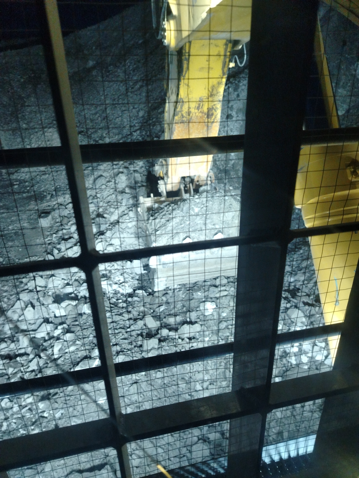The image shows a view from above, through a metal grate, of a mining operation. The ground is covered with rubble and rocks, and a yellow piece of heavy machinery, likely a loader or excavator, is visible. The machine appears to be in operation, as there are indications of disturbed earth and debris. The scene is dimly lit, suggesting it is either night or underground. The overall impression is one of an active industrial site. The specific location in Naminga, Russia, is not depicted in the image itself.