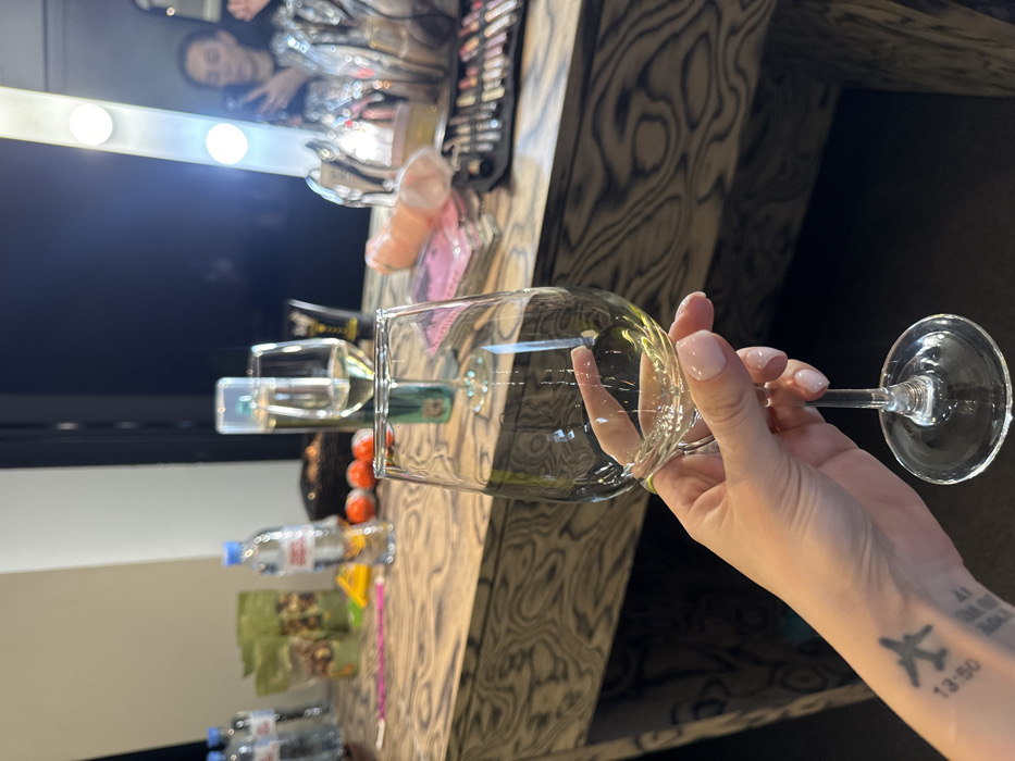 A female's left hand, featuring a silver ring on the ring finger and a wrist tattoo depicting an airplane with the text "13:50" and additional partial text, holds a clear stemmed wine glass containing a light yellow liquid. The hand has light pink manicured fingernails.
The hand is positioned in front of a wooden-patterned table. On the table, from left to right, are two plastic bottles of Evian mineral water, two green foil-like bags, a dark basket containing indeterminate items, three orange Kinder Surprise eggs, another stemmed wine glass with light yellow liquid, a tall clear bottle with a green label, a black cosmetic tube, a pink phallic-shaped object, a stack of pink rectangular items, and an open black makeup case filled with various brushes and cosmetic tools.
In the background, a large vanity mirror with exposed light bulbs along its frame reflects a female individual. This individual, partially obscured, is holding a mobile phone and appears to be wearing a shiny, possibly sequined, garment. The room behind the mirror reflection is dark, while walls in other areas of the scene appear light-colored.
The specific location within the city of Moscow, Russia, is not discernible from the visual information provided. The scene is an indoor setting, likely a dressing room or preparation area.