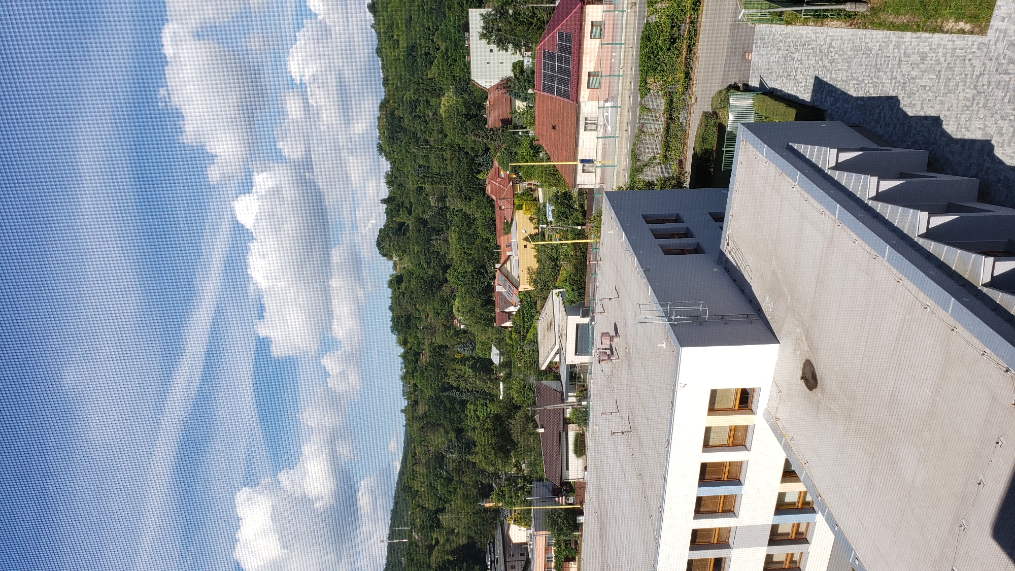 The image depicts a view from a window covered by a screen, looking out onto a suburban area. In the foreground, the roof of a modern, light-colored building is visible, characterized by its flat surface and several ventilation pipes. Adjacent to this building is another section of a building with a similar flat roof.
Beyond these structures, a cluster of residential houses with pitched roofs, varying in color from red to yellow and brown, are nestled amongst lush green trees. The houses appear to be part of a well-established neighborhood with paved roads and sidewalks. In the distance, a tree-covered hillside rises, indicating a natural landscape surrounding the urban development. The sky is a clear blue with scattered white clouds.
This scene is consistent with a residential area in the city of Prešov, Slovakia, likely on the outskirts where the urban environment meets a more natural, hilly terrain. There are no people visible in the image.
