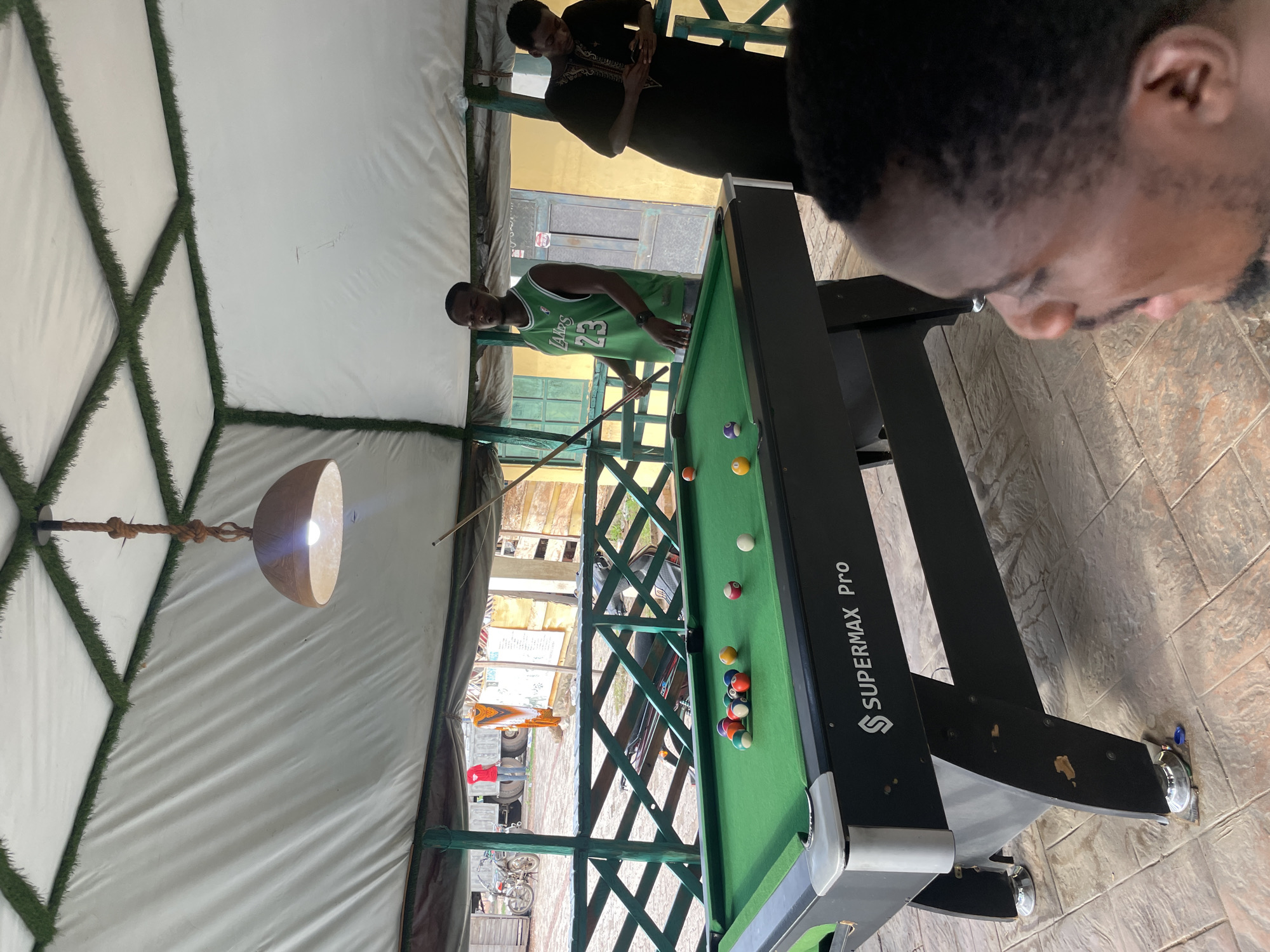 The image depicts a scene indoors, seemingly under a tent-like structure. The location is identified as Lokoja, Nigeria.
There is a pool table dominating the space. A man wearing a green Lakers jersey with the number 23 is poised to strike a billiard ball. Another individual, wearing a dark patterned shirt, is standing near the table observing the game. A third person's head is visible in the right side of the photo.
The pool table has a green surface and features a "SUPERMAX Pro" logo. Several billiard balls are scattered on the table. A light hangs above the pool table illuminating the scene.
The surroundings appear to be a partially enclosed space. Some outdoor elements, such as fencing and what looks like a vehicle, are visible through openings. The flooring looks like tile.