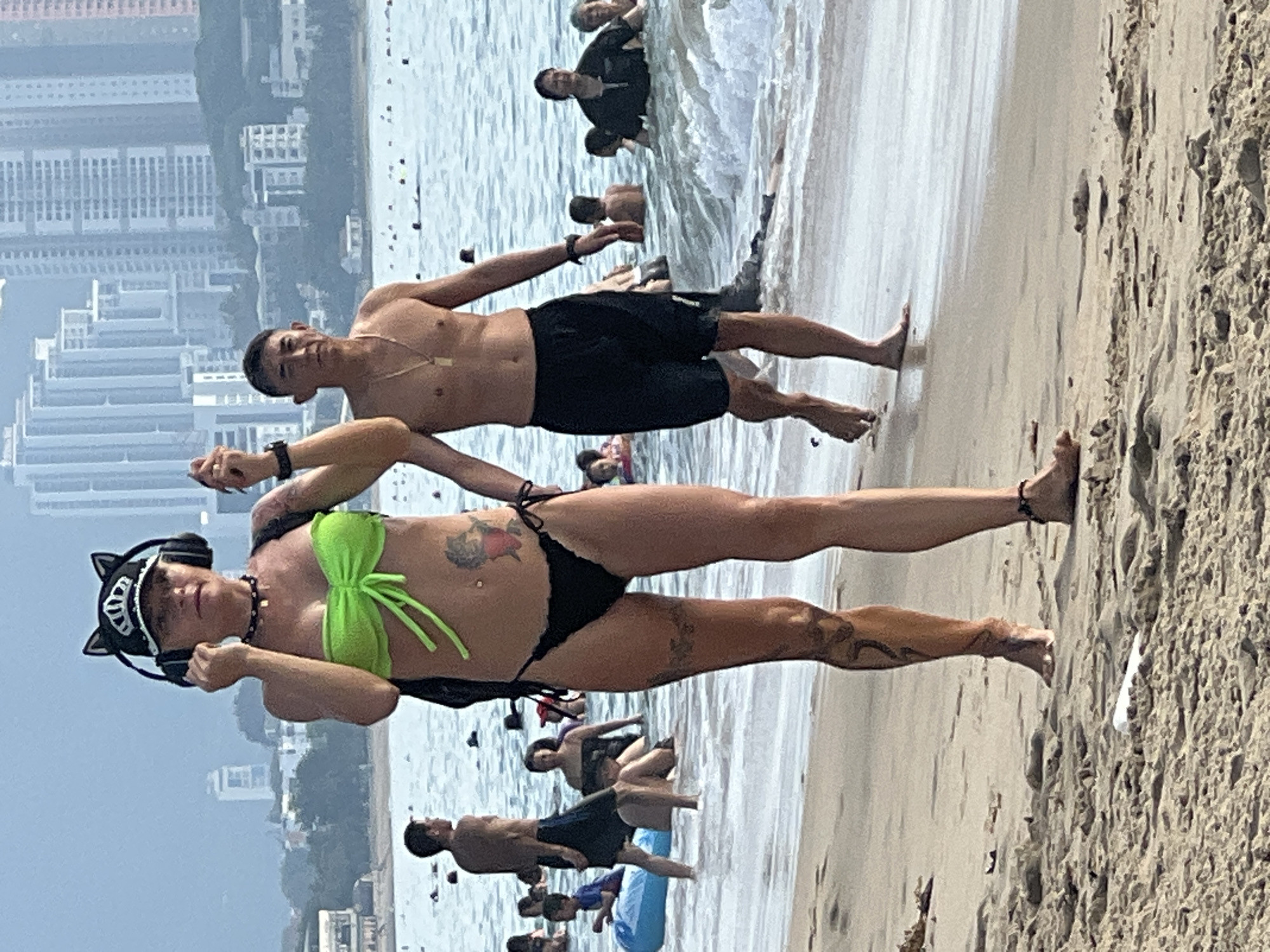 In Nha Trang, Vietnam, a woman wearing a black cap with cat ears and headphones stands on a sandy beach, her body angled to the left. She is dressed in a bright green bikini top and a black bikini bottom. Her midsection displays tattoos, and her legs are also tattooed. A man in black shorts and without a shirt walks to her right and slightly ahead, facing forward. In the background, the ocean is filled with people swimming and wading. Beyond the water, a cityscape with tall buildings is visible. The scene depicts a typical day at the beach with recreational activities and urban infrastructure.