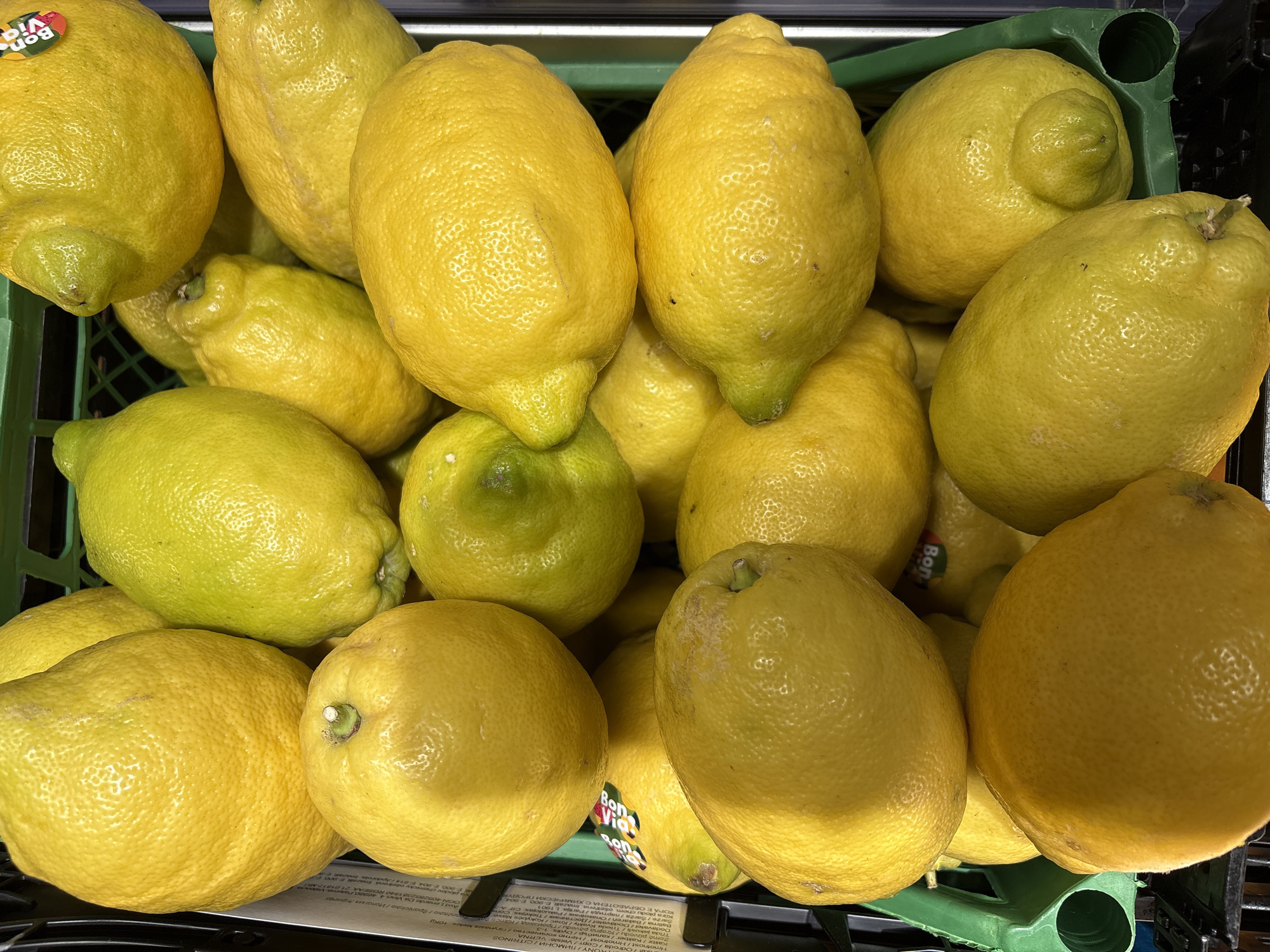 A close-up, high-angle shot reveals a green plastic crate overflowing with unwaxed lemons of varying sizes and colors, predominantly yellow with some tinges of green. Some lemons bear stickers, notably one with the "Bon Vida" logo. The lemons appear to be at a market or grocery store, with part of a printed label visible beneath them. The label includes information in several languages, including Bulgarian. The text in the OCR includes "Лимони" which translates to "Lemons" in Bulgarian. It is not possible to determine that this image was taken in the city of Varna, Bulgaria.
