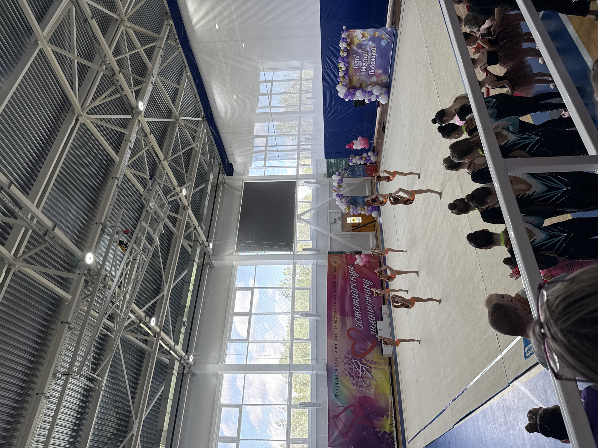 Here is a detailed description of the image:
**Location:** The image appears to be taken at an indoor sporting event, possibly a rhythmic gymnastics competition. The text on the banner hints this is in the city of Veliky Novgorod, Russia.
**Environment:** The setting is a large gymnasium or sports hall with a high ceiling supported by a steel structural framework. Large windows provide natural light, and there is a banner decorated with balloons and text written in Russian.
**People:** There are several groups of people visible. On the left, a group of athletes in matching uniforms (likely rhythmic gymnasts) stand on a small podium. On the right, there are spectators standing behind a barrier, watching the event. The image is taken from the perspective of a spectator.
**Objects:** Notable objects include the large structural framework supporting the roof, the windows, decorative banners, and the podium the athletes are standing on. The floor appears to be a light-colored, matted surface.
**Actions:** The athletes on the podium are likely receiving recognition or awards. The spectators are watching the event.
**Interactions:** The athletes are likely interacting with the event organizers. The spectators are focused on the athletes and the event taking place.