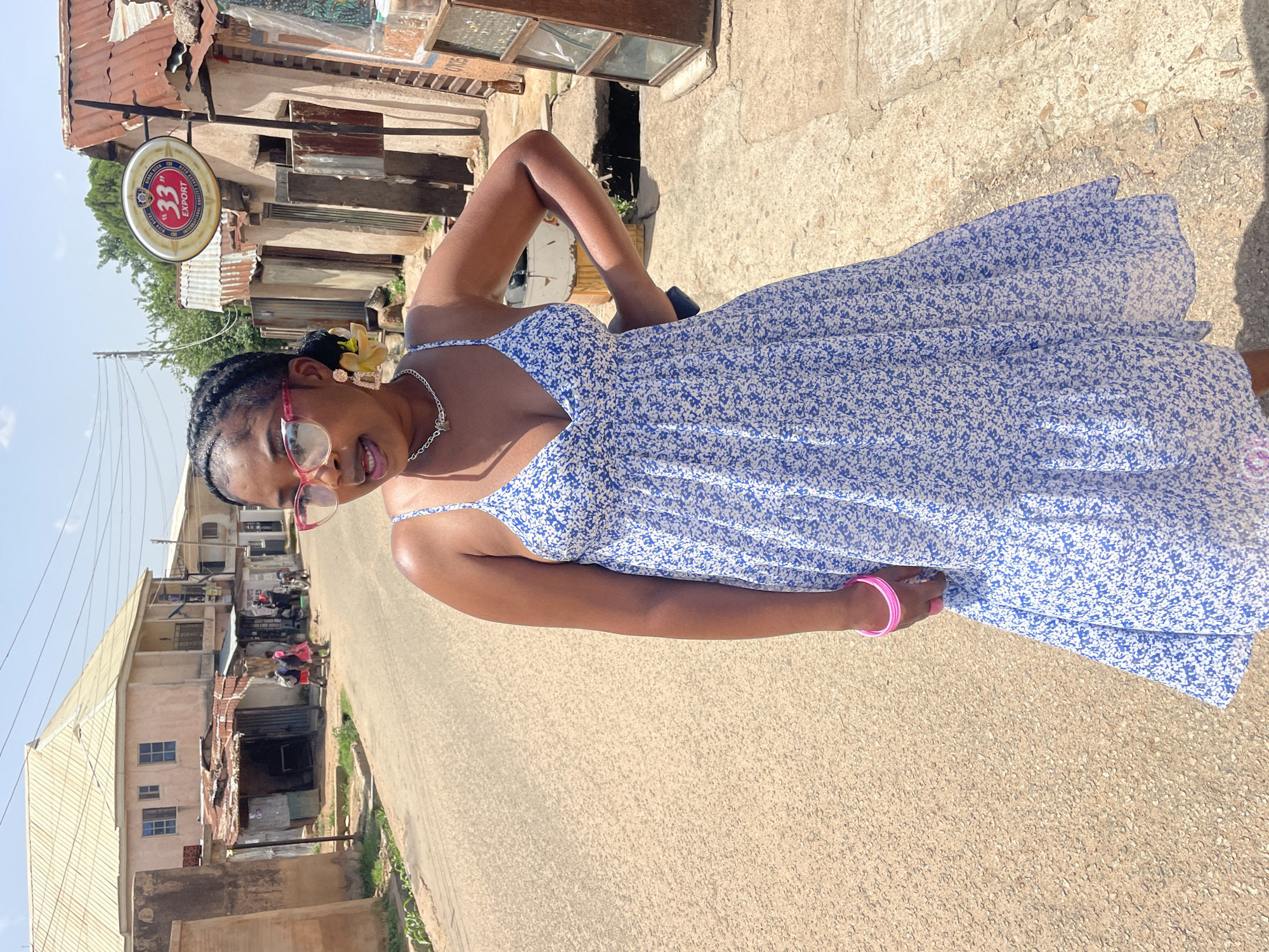 A person is posing for a photo outdoors, standing on a paved road. They are wearing a blue and white floral print dress, pink-rimmed glasses, and a necklace. A yellow flower is tucked into their hair. The person is looking towards the camera with a slight smile.
In the background, there are buildings with corrugated metal roofs, typical of a marketplace or informal settlement. A sign for "33 EXPORT" beer is visible. Several motorcycles and people can be seen further down the road, suggesting a bustling environment. The scene appears to be in Jos, Nigeria, during the daytime, under clear skies. The lighting suggests it is a sunny day. The overall mood is casual and relaxed, with the subject appearing confident and happy.