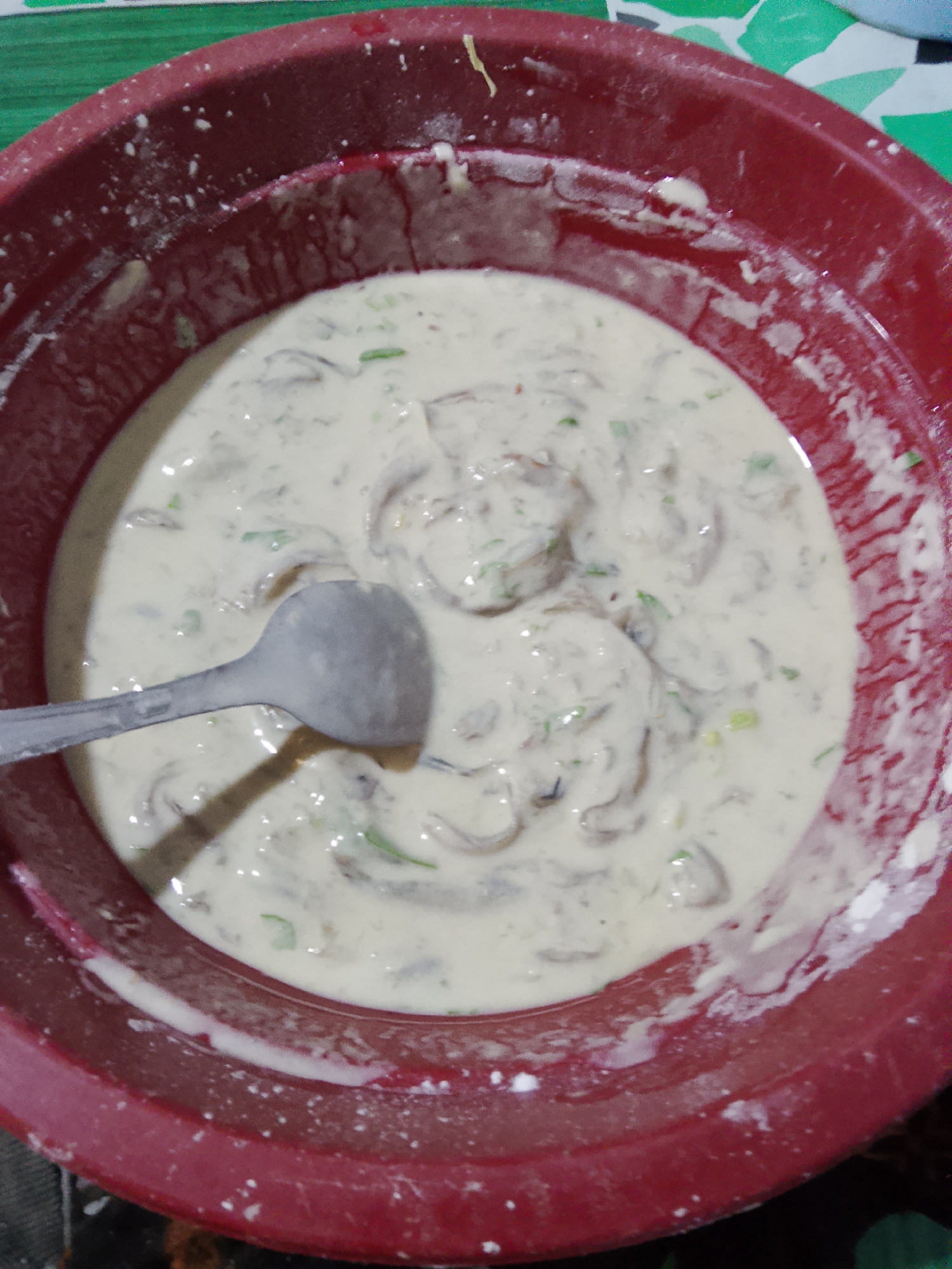 A close-up, overhead shot shows a red plastic bowl filled with a creamy batter containing thinly sliced ingredients. A silver spoon rests within the bowl, partially submerged in the mixture. The batter is a light beige color, with visible pieces of what appear to be mushrooms and green herbs dispersed throughout. The rim of the bowl is also coated with some of the batter, and there are white specks, possibly flour, scattered on the interior and exterior. The background is out of focus, but suggests a green surface, possibly a tablecloth, with a geometric pattern. The image does not provide any information that would allow for a specific location within the city of Merbopuntong, Indonesia to be identified.