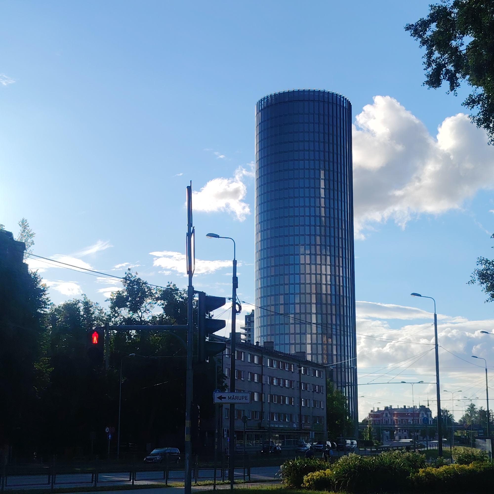 This image captures a bright, sunny day in Riga, Latvia, with a clear blue sky dotted with fluffy white clouds. The primary subject is a modern, cylindrical skyscraper with a glass facade that reflects the sky. To the left of the skyscraper, a multi-story gray building with many windows is visible. In the foreground, a traffic light shows a red pedestrian signal, indicating that people should not cross. A street sign with an arrow pointing left reads "MĀRUPE," which is likely a direction to a location or district. Trees and greenery are abundant throughout the scene, framing the urban landscape. The overall atmosphere is calm and urban, with a sense of everyday life unfolding. The time of day appears to be afternoon, given the sunlight and shadows.