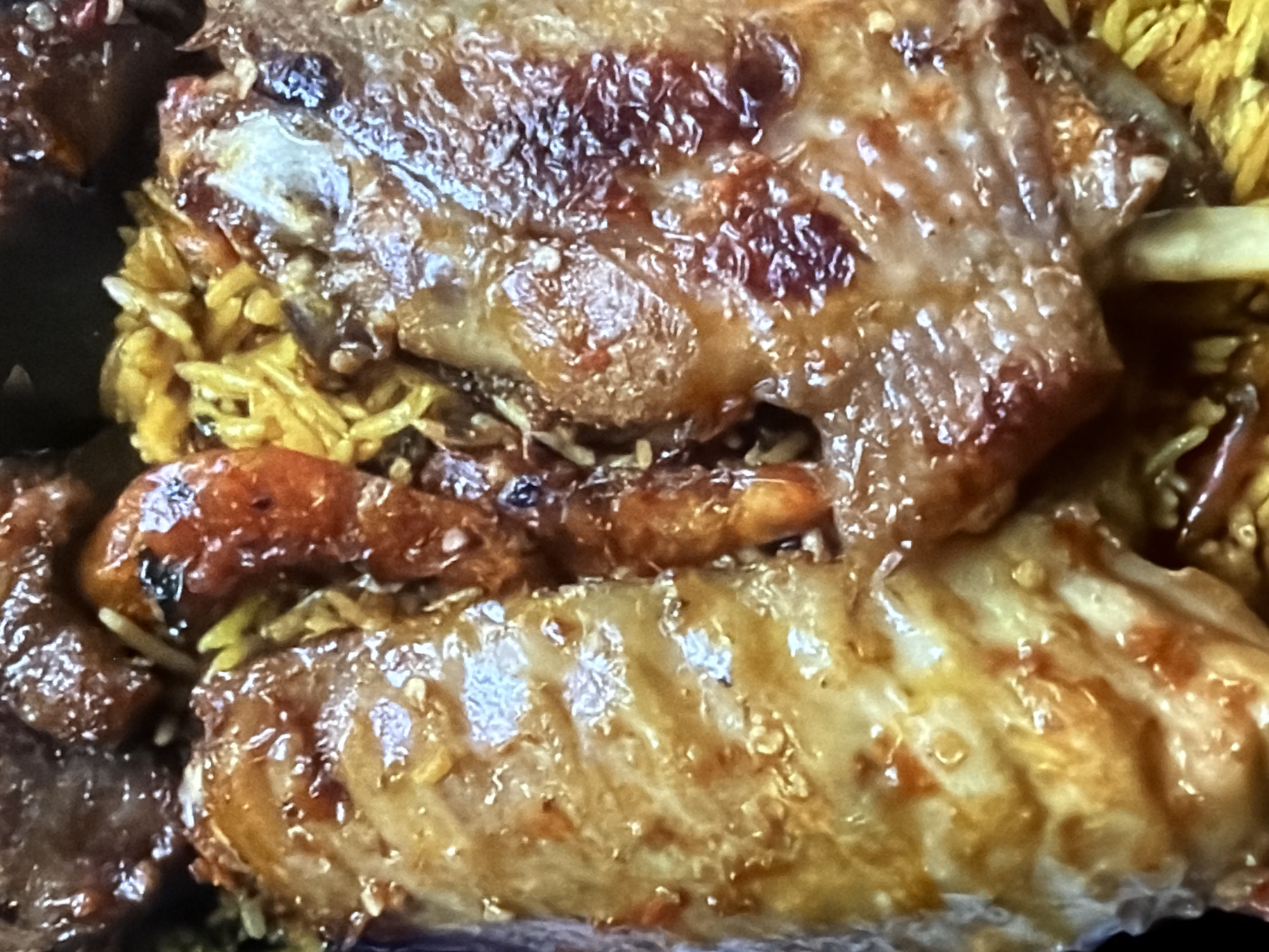 The image displays a close-up, overhead view of a serving of food, likely a meat dish with rice. The food is presented in a dark container or plate. Two large pieces of meat, appearing to be braised or roasted, are prominent. One piece on the left shows layers of meat and bone structure, glistening with a rich, amber-colored sauce that has flecks of red and orange, possibly from spices or a glaze. The meat itself has a light beige to golden-brown color, with some darker, caramelized areas.
To the right of the first meat piece, and slightly above it, is another substantial piece of meat, also coated in the glossy sauce. This piece exhibits deeper reddish-brown hues in certain areas, suggesting more intense caramelization or a different cut. A bone protrudes from the bottom of this piece.
Interspersed between and around the meat is a bed of rice, appearing to be a pilaf or seasoned rice, with a distinct yellow-orange color. There are also hints of darker ingredients mixed within the rice, such as small pieces of meat or vegetables. In the upper part of the image, more pieces of dark, rich-looking meat are visible, partially obscuring the background. The lighting is direct, creating highlights on the glossy surfaces of the food.
The context provided places this scene in the city of Bukuru, Nigeria. Without further information, it is not possible to determine specific locations within Bukuru. The image is purely a food presentation, with no visible people, actions, or environmental details beyond the food itself and its immediate serving container.