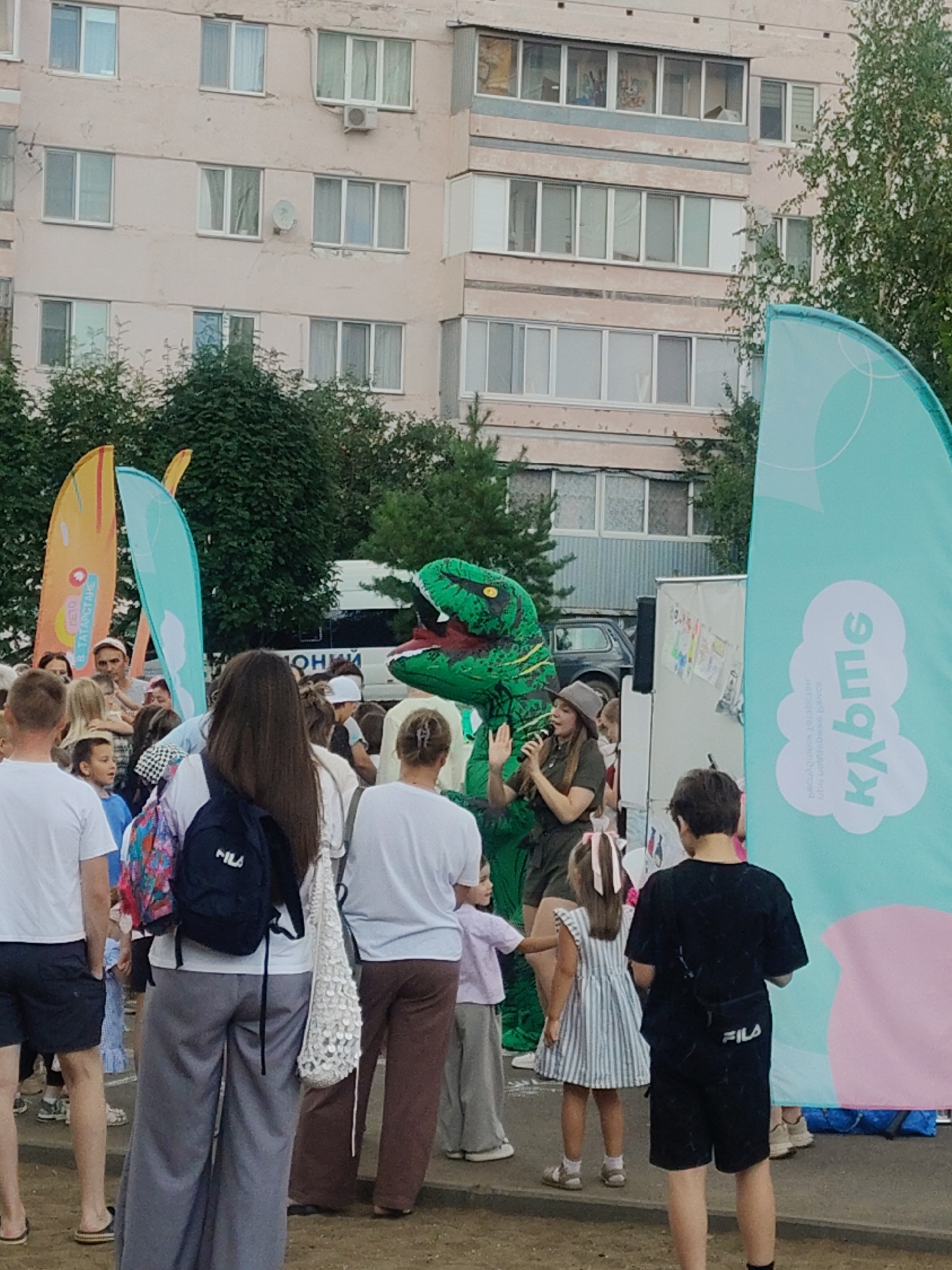 An outdoor event is taking place in front of a multi-story apartment building in Mendeleyevsk, Russia. The building has pale pink siding and numerous windows. In the foreground, a crowd of people, including adults and children, are gathered. To the left, two large, brightly colored banners are visible, one orange and one turquoise. In the center of the scene, a person in a green dinosaur costume is interacting with a woman holding a microphone. The woman is wearing a hat and a green outfit. A stage or platform is behind them, with some children's drawings displayed on it. To the right, a large turquoise banner with white lettering and cloud graphics is prominently featured. Several people are facing the stage area, appearing to watch the performance. The ground is a mixture of pavement and dirt. Trees and foliage are visible in the background.