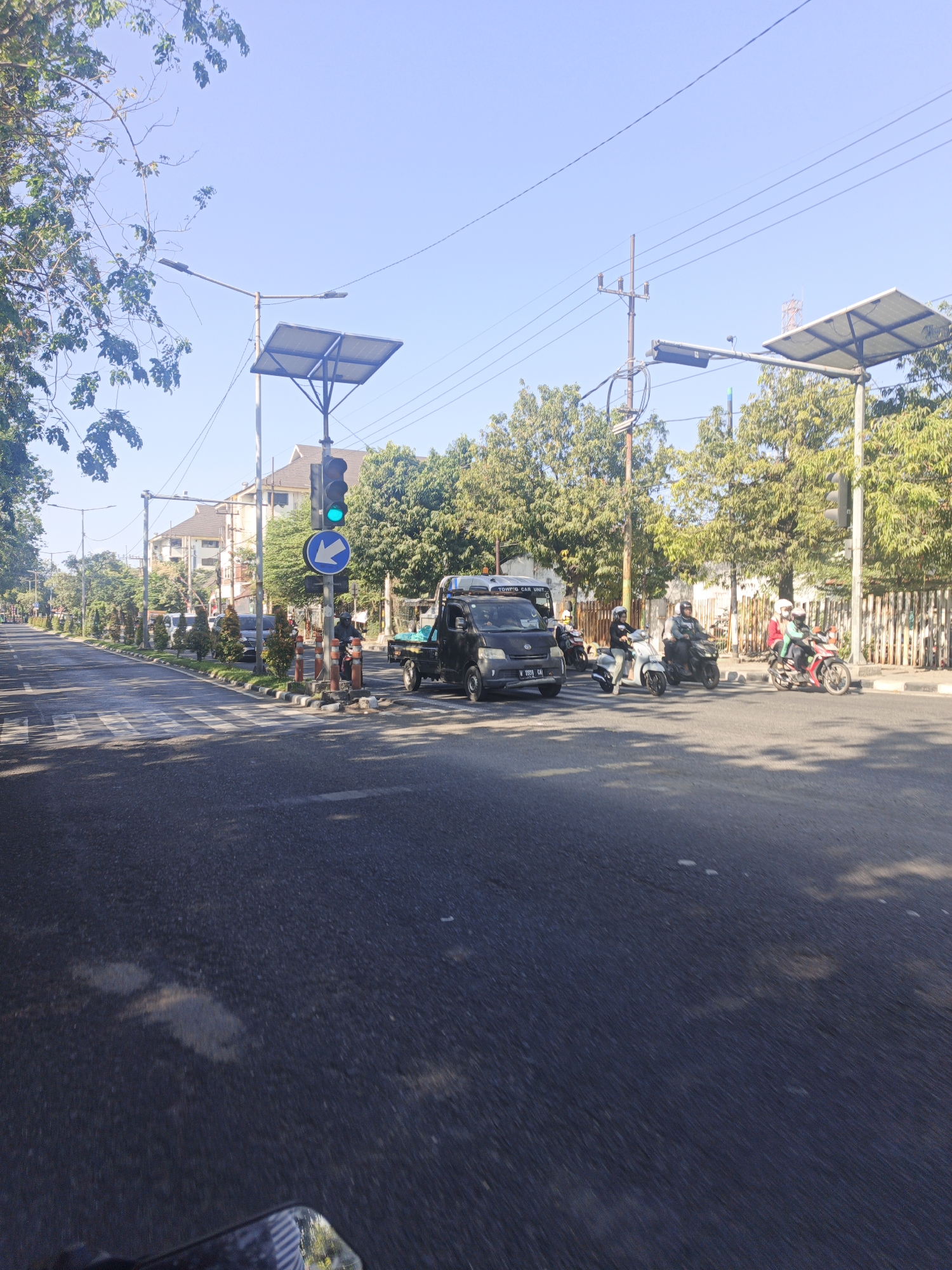 The image depicts a street scene in Surabaya, Indonesia, likely during daylight hours given the bright sun and clear blue sky. The foreground shows an asphalt road with a pedestrian crossing. A traffic light with a green signal is prominently displayed, along with a blue directional sign indicating a left turn. Several vehicles are present at the intersection, including a black truck, multiple motorcycles with riders, and a dark-colored van. The surrounding environment includes trees lining the street, buildings in the background, and utility poles with power lines overhead. Solar panels are mounted on poles near the traffic lights. The overall impression is that of a typical urban intersection with moderate traffic activity.