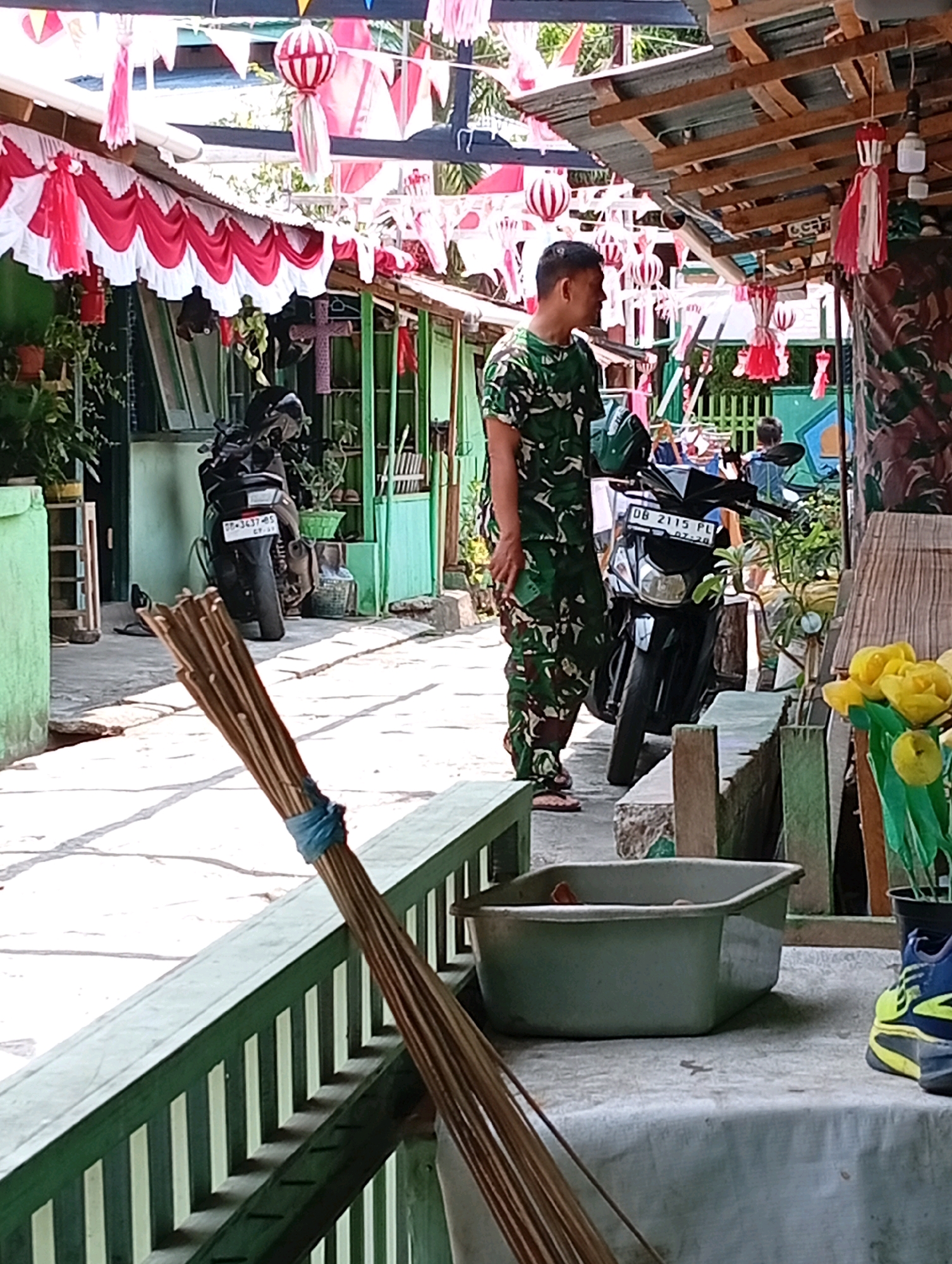 A man in camouflage uniform stands near a black motorcycle. He is looking to his left. The scene appears to be outdoors, possibly a street or courtyard, with buildings and a wooden roof structure visible. Red and white decorations, including flags and lanterns, are hung overhead, suggesting a festive or patriotic occasion. In the foreground, a broom made of straw lies on a green wooden railing. To the right, yellow flowers in a pot and a blue and yellow shoe are also present. The overall environment suggests a tropical or warm climate, with bright sunlight casting shadows. The license plate on the motorcycle reads "DB 2115 PL". The presence of military-style camouflage attire and the decorations might indicate a connection to Indonesian national holidays or events. Given the context, this scene could be in Manado, Indonesia, a city known for its vibrant cultural celebrations and military presence.