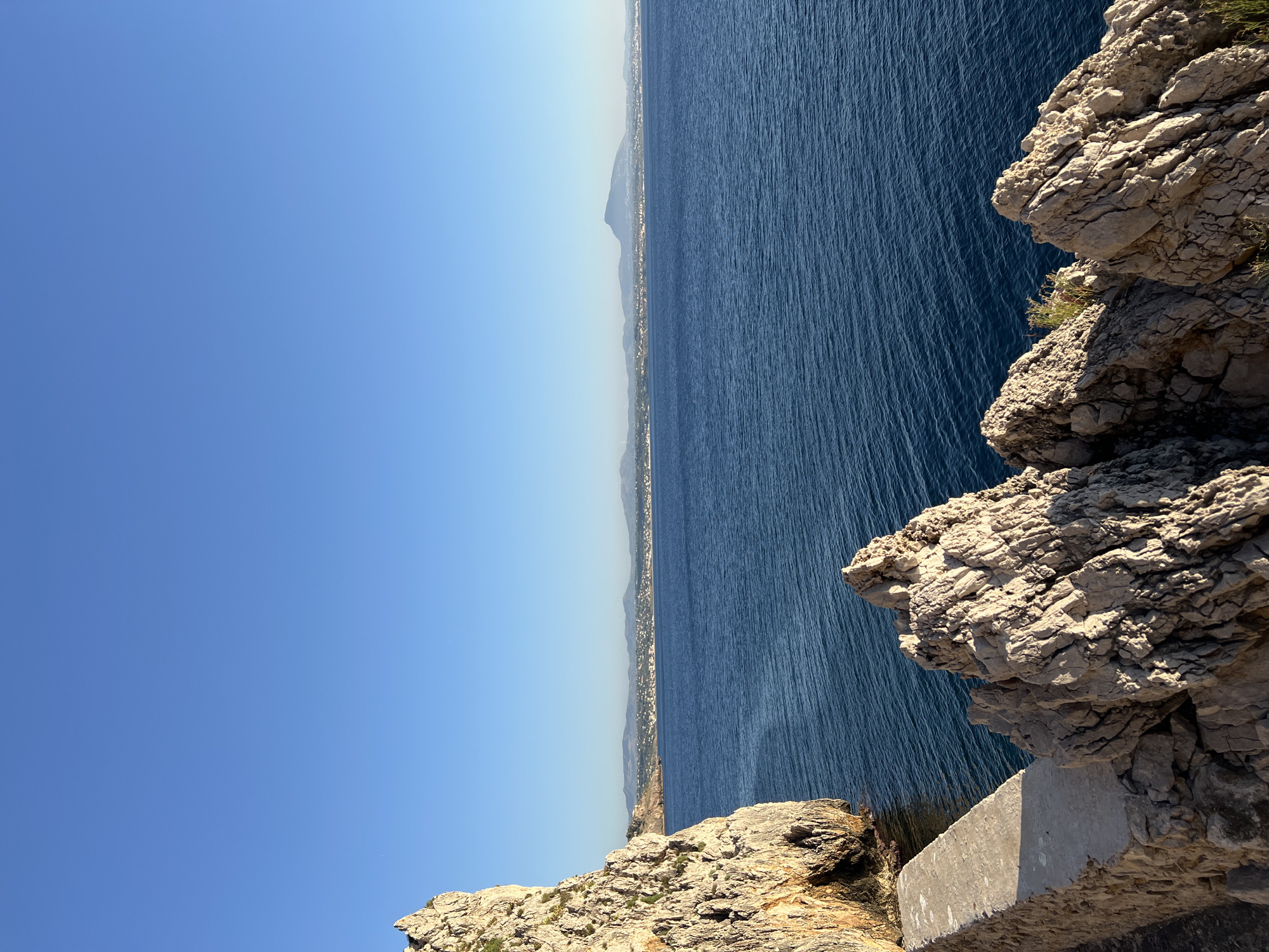 The image depicts a serene coastal landscape under a clear blue sky. In the foreground, rugged, light-colored rocks dominate, with some exhibiting sharp, angular formations. A concrete structure, possibly a walkway or retaining wall, is visible along the left side of the foreground. Beyond the rocks and the concrete structure, a calm, deep blue sea stretches out to the horizon. The distant shoreline features a town with buildings and is backed by rolling hills and mountains. The sun is casting shadows, indicating it is likely morning or late afternoon. This scene is suggestive of a location along the coast of Terrasini, Italy, known for its picturesque rocky shores and clear Mediterranean waters. There are no people or animals visible in the image.