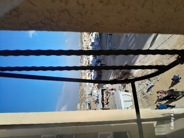 The image depicts a view from a high vantage point, possibly a balcony. A railing partially obscures the scene below, which appears to be a densely populated area with makeshift shelters or buildings. People are visible on the ground. The sky is visible in the upper left portion of the frame. A building or structure frames the top and bottom of the image. The overall impression is of an impoverished or developing urban environment.
