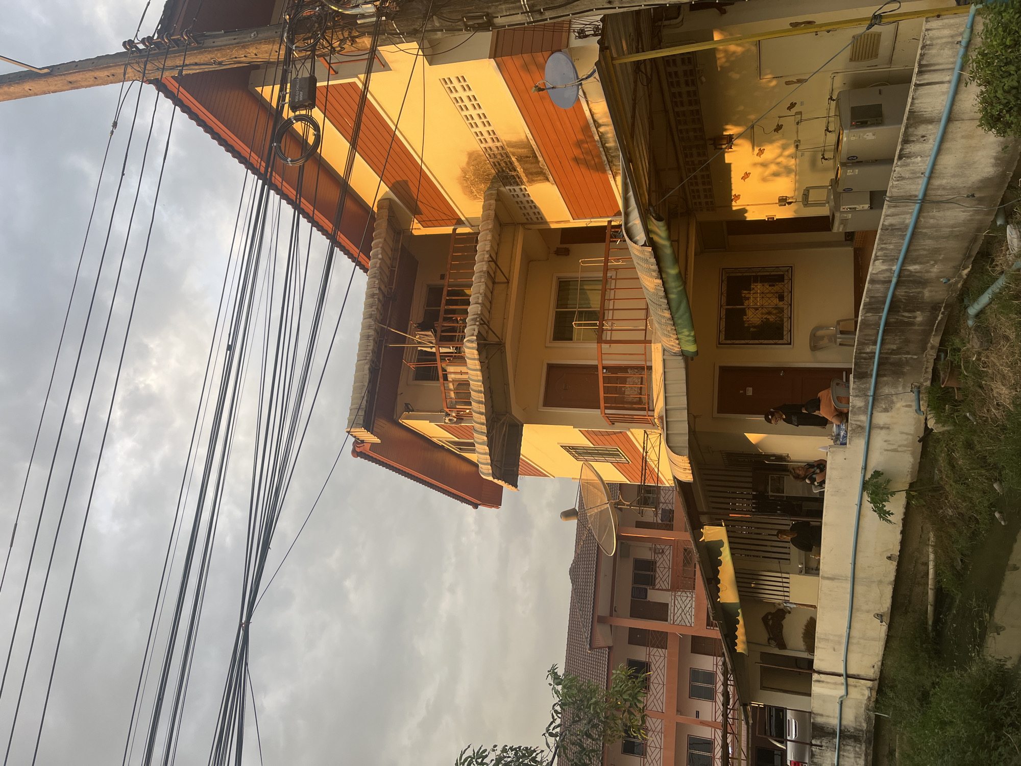 The image depicts a street scene in บ้านกาญจน์กนก 23, Thailand. A two-story building with a yellow and red striped facade dominates the view, featuring balconies with metal railings and a satellite dish on the roof. Electrical wires are heavily layered against a cloudy sky. Adjacent to this building is a concrete wall with a blue pipe running along it. On the ground level of the building, several people are gathered on a porch or covered area. Another building, with a similar architectural style, is partially visible in the background, featuring an orange and white facade. A car is parked in front of the second building. Vegetation is present near the base of the second building.