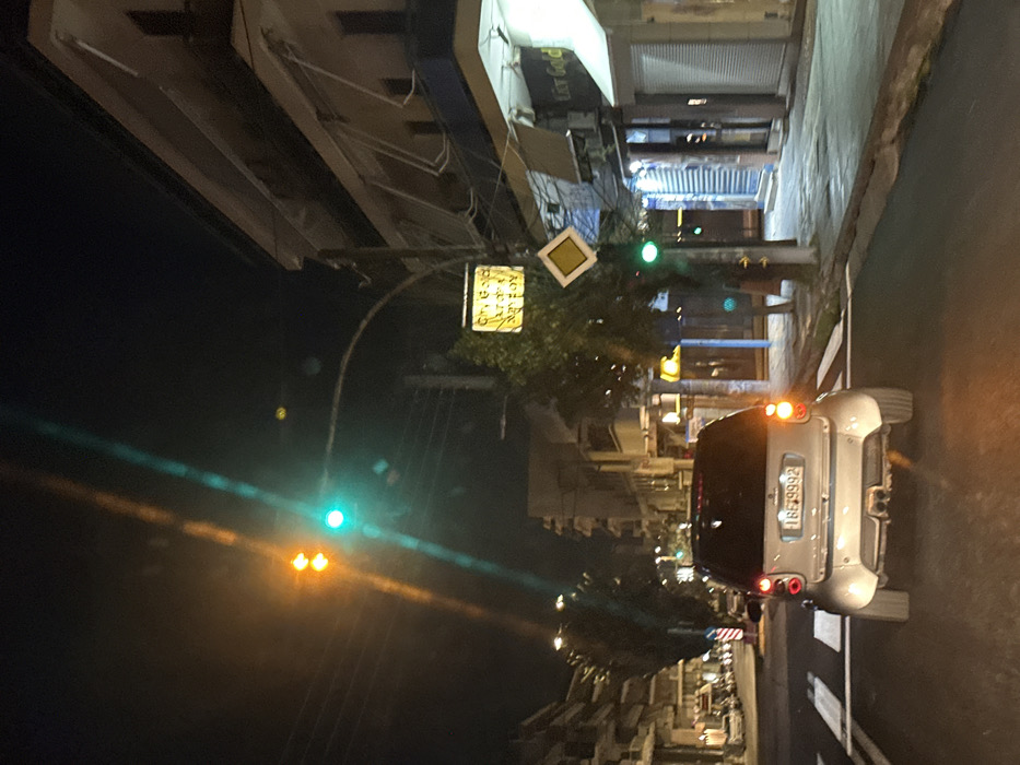 The image depicts a nighttime street scene in a city, likely Iraklio, Greece, viewed from a slightly elevated perspective. The street runs along the right side of the frame, bordered by a sidewalk and buildings on the left.
In the foreground on the right, a silver compact car, possibly a Smart car, is parked or stopped with its red taillights illuminated. Its Greek license plate, "IBΓ 9992," is clearly visible on the rear. The street features white markings.
Along the left side, several multi-story buildings line the street. Above them, hanging over the roadway, are two prominent signs. One is a yellow diamond-shaped sign, indicating a priority road in European traffic signage. Below and to its left, a rectangular yellow sign with indistinct Greek text is visible, possibly advertising a local business. Another faint, blurry sign on one of the building awnings appears to say "Eat Good". A street lamp arches into the scene from the left, with visible green and yellow traffic lights.
The time of day is clearly night, with the sky being dark and artificial lights from street lamps, traffic signals, and buildings providing illumination. The atmosphere appears calm, and no people are visible. The presence of Greek text and a Greek license plate strongly indicates the location context of Iraklio, Greece.