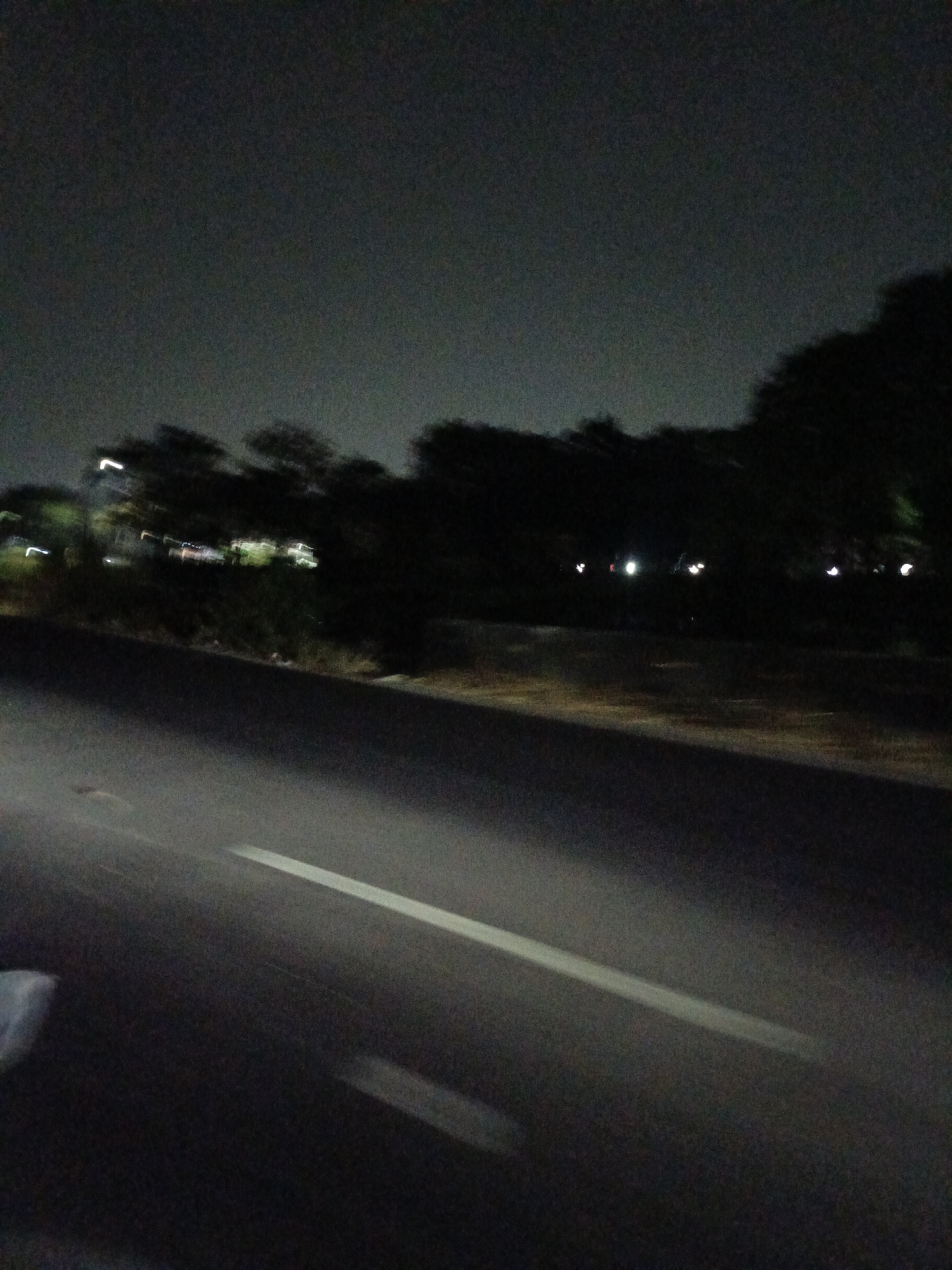 The image shows a road at night, likely in Gandasari, Indonesia. The road surface is dark asphalt with a white line marking. Trees and foliage are visible on the right side of the road, silhouetted against a dark sky. There are some blurred lights in the background, suggesting buildings or streetlights. The overall impression is of a quiet, possibly rural or suburban road at night.