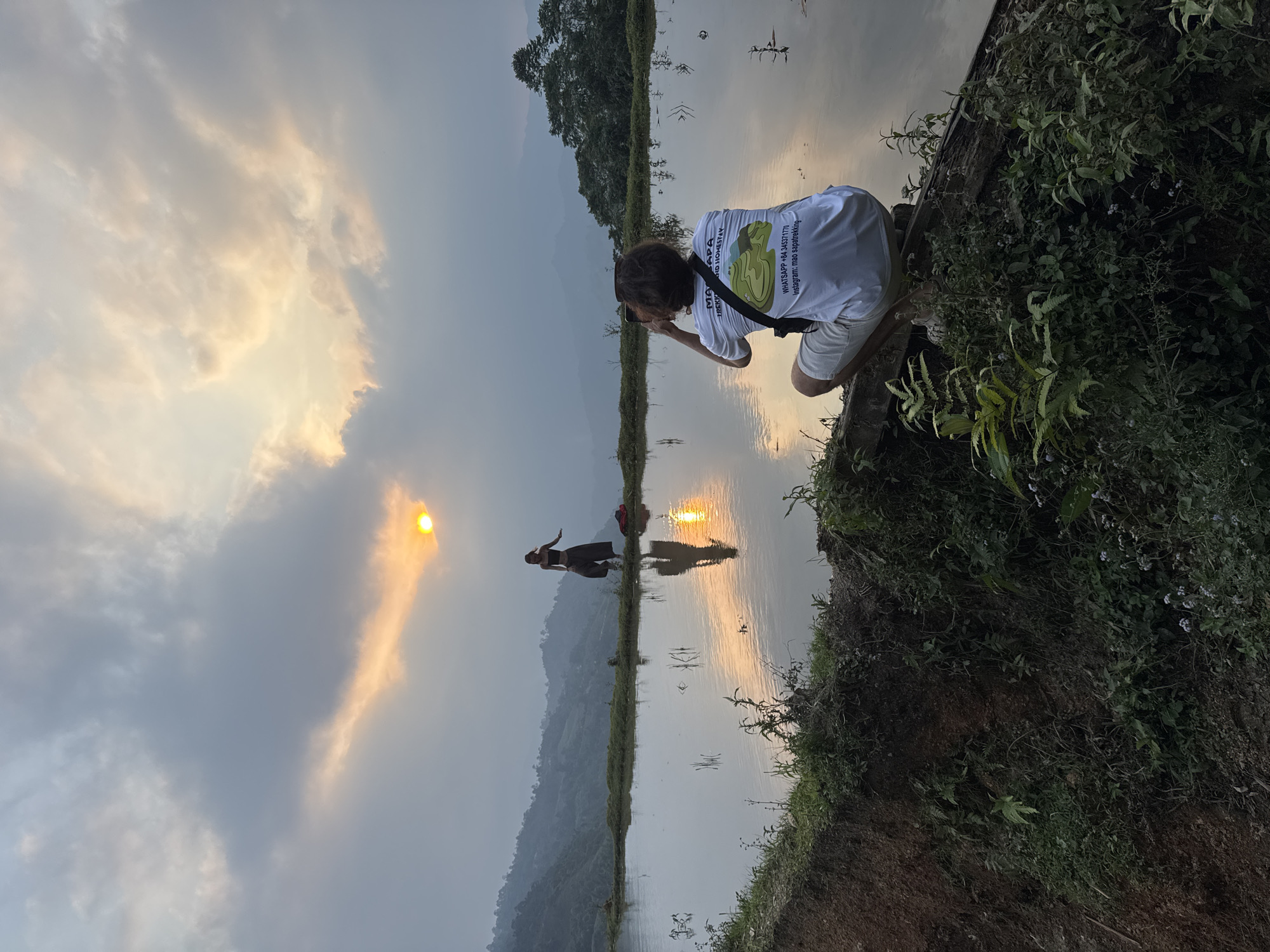 The image depicts a serene sunset scene over water-filled rice paddies in Nậm Dịch Commune, Vietnam. The sky is partly cloudy, with the setting sun visible as a bright golden orb in the upper left, casting a warm, orange glow across the clouds and the landscape.
In the foreground, a person with dark, medium-length hair is crouched on a grassy bank on the right, holding a smartphone to capture the view. They are wearing a white t-shirt and light-colored shorts. Visible text on their t-shirt reads "MAPA", "M&D HOMESTAY", and contact information: "WHATSAPP: +84 343717779", "instagram: mao.sapatrekking", accompanied by a graphic depicting mountains.
Further back, another individual stands on a narrow, elevated path that separates two sections of the water-filled paddies. This person is largely silhouetted against the brilliant reflection of the sun on the water, which creates an elongated, golden streak across the surface. A red backpack is visible near them on the path. The person appears to be posing or simply admiring the scenic sunset.
The tranquil water acts as a mirror, reflecting the vibrant sky and the sun's glow. Distant hills or mountains form a hazy backdrop, adding depth to the vast landscape. Lush green vegetation, including ferns and other plants, grows on the banks bordering the water, enhancing the natural beauty of the rural setting. The overall atmosphere is peaceful, capturing a moment of appreciation for nature at dusk.