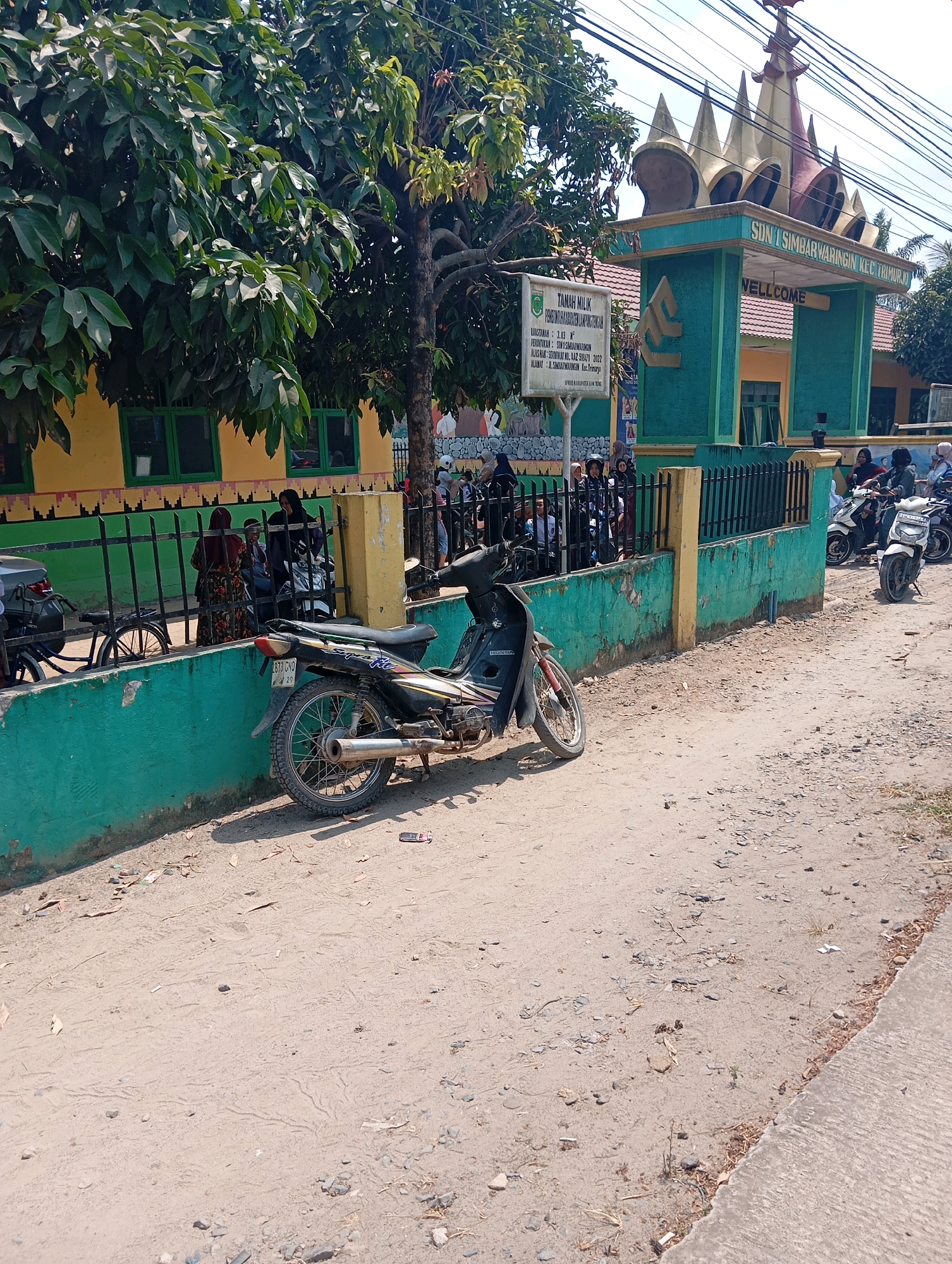 The image captures a street scene outside a school building in Batang Hari Ogan, Indonesia. The school's entrance gate is prominent, featuring a decorative, crown-like structure at the top and teal-colored pillars. Text on the gate reads "SDN 1 SIMBARWARINGIN KEC. TRIMURJO" and "WELCOME." A sign to the left of the gate displays information about the land, indicating it is owned by the government and has an area of 3.83 hectares.
In front of the school, a black motorcycle is parked on a dirt and gravel road. The road is uneven and appears to be unpaved. To the left of the motorcycle, a green wall runs along the school's perimeter, with a black metal fence atop it. Behind the fence, several people, mostly women and children, are gathered. Some are wearing hijabs. A bicycle is also visible behind the fence. In the background, trees with dense green foliage provide shade. Further down the road, more motorcycles are parked, and people are seen interacting. The overall environment suggests a sunny day, with shadows cast on the ground. The building itself has yellow walls and green window frames.