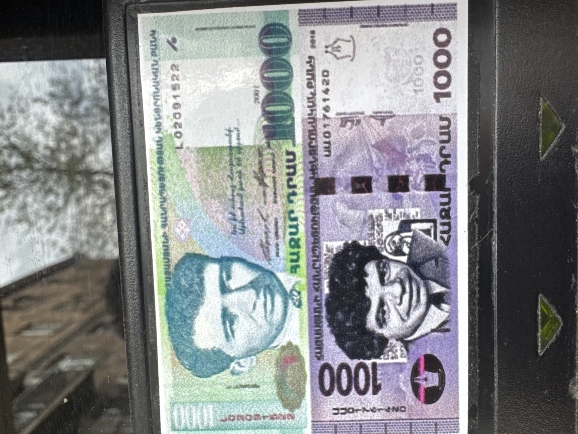 The image depicts a 1000 Armenian dram banknote. The note is visible partially due to its proximity to a window and part of a device with directional arrows. The background is slightly visible due to the reflection of the window. The banknote shows various details including portraits, serial numbers, and the denomination "1000" written in both numbers and Armenian script. It features different shades of green and purple. It does not give any information about the location of Yerevan or Armenia.