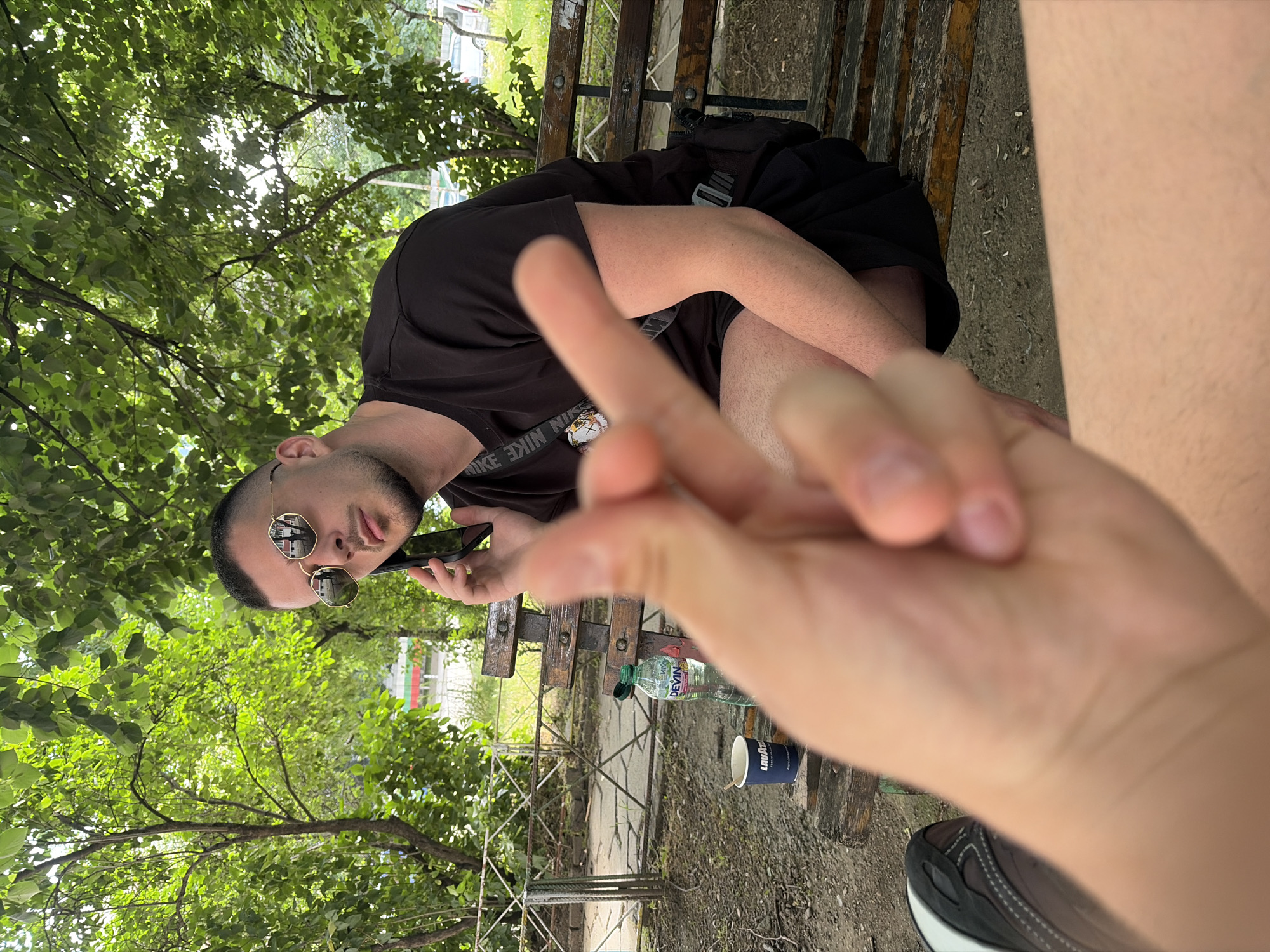 The image captures an outdoor scene in a park or urban green space, identified as being in Sofia, Bulgaria, due to a visible red, white, and green striped national flag on a distant building. In the immediate foreground, a light-skinned hand is out of focus, performing a gesture where the index finger and thumb form a loop. Part of a black and white sneaker is visible on the lower-left. In the mid-ground, a light-skinned male with short dark hair, a beard, and gold-rimmed mirrored sunglasses is seated on a rustic wooden bench. He wears a black t-shirt, black shorts, and a black cross-body bag with "NIKE" branding. He is holding a dark-colored smartphone to his right ear, appearing to be engaged in a phone conversation. On the bench beside him are a partially full "DEVIN" brand mineral water bottle with a green cap and a blue "LAVAZZA" branded paper coffee cup containing a stirrer. The setting is characterized by numerous green trees and foliage, with sunlight filtering through the leaves. The ground is composed of dirt and gravel, and a low metal fence is visible behind the bench, separating the immediate area from the distant background which includes indistinct vehicles and structures.