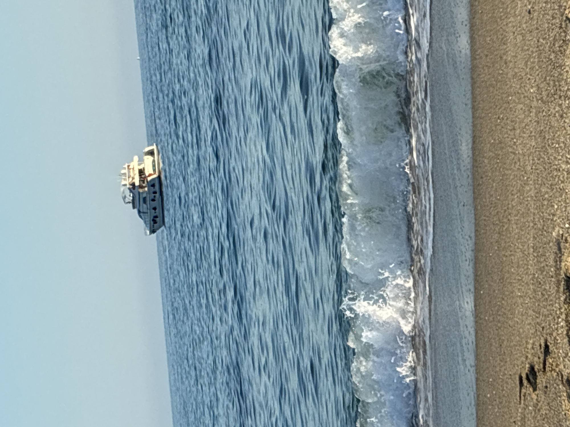 The image depicts a bright, sunny day at a beach in Alanya, Turkey. The foreground shows a sandy beach with some faint footprints and a small wave breaking at the shore. The middle ground is dominated by the blue, rippling sea, with a white-capped wave prominently featured. In the distance, a white yacht is sailing on the calm sea under a clear blue sky. The overall scene evokes a peaceful and scenic coastal environment.