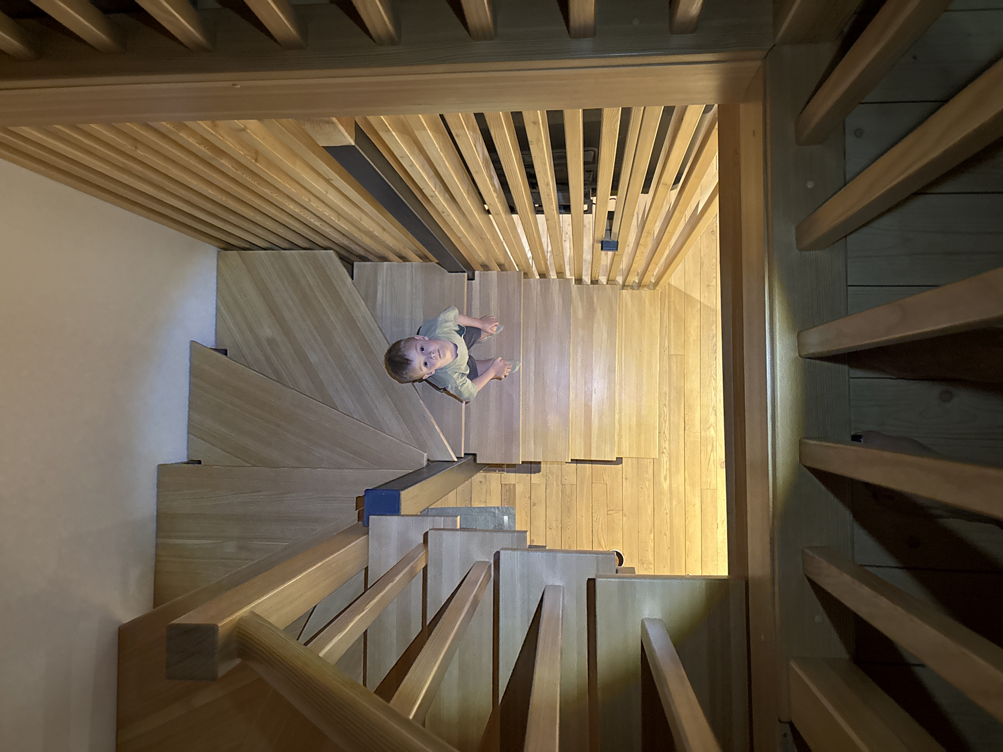 A high-angle, top-down shot captures a young child looking up from a multi-level wooden staircase in a modern interior, located in Chaikovsky, Russia. The child, a boy, stands on a landing or wide step, dressed in a light-colored t-shirt and shorts, gazing directly upwards towards the camera with a curious expression.
The architectural space is characterized by extensive use of light-colored wood, possibly oak or a similar species. This material forms the entirety of the staircase, including its treads, risers, and the robust wooden balustrade composed of numerous vertical slats. The staircase design is contemporary, featuring sharp angles and a geometric, almost sculptural aesthetic for the steps and railings. Wooden planks also make up the visible floor sections at various levels.
Above the child, a distinctive slatted wooden ceiling or upper wall structure is visible, mirroring the vertical lines of the balustrade. Integrated into this structure and within the stair railings are some darker, possibly metallic, accents. Sections of plain white wall provide a neutral backdrop against the dominant wooden elements. The scene is brightly and uniformly illuminated, likely by indoor artificial lighting, which highlights the warm tones and textures of the wood and creates subtle shadows that define the architectural forms. No visible text is present in the image.