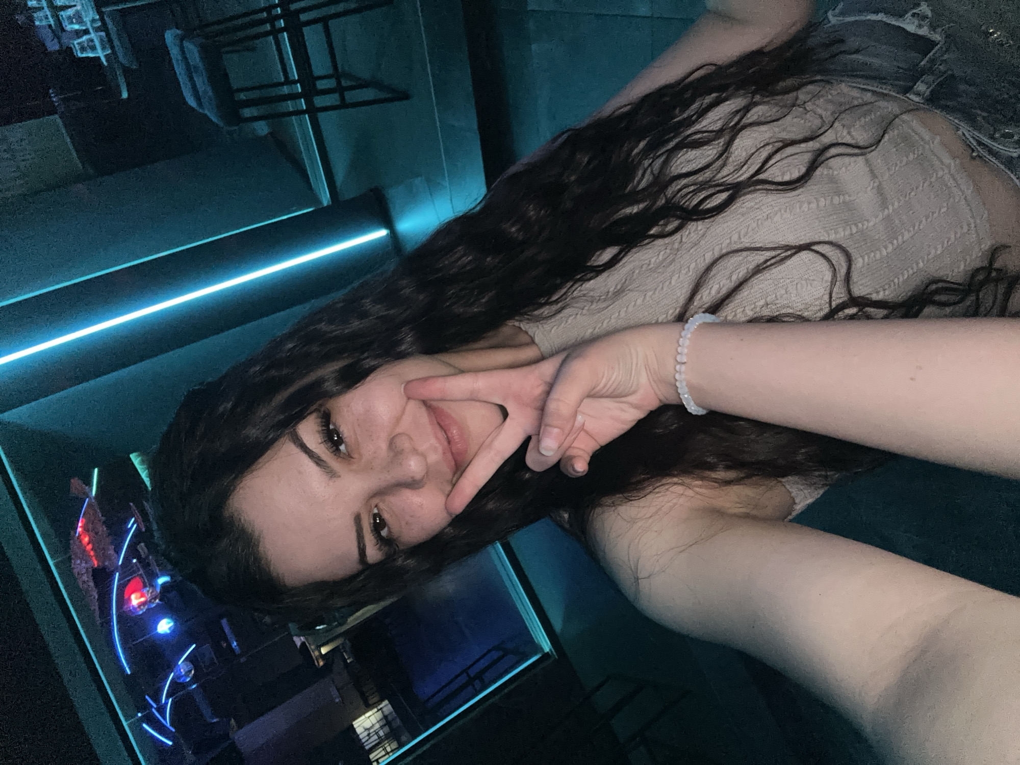 A young woman is depicted taking a selfie indoors in a dimly lit establishment within the city of Sliven, Bulgaria, likely a bar or club. She has long, dark, wavy hair and is smiling subtly with her lips closed. Her left hand (viewer's right) is raised, forming a peace sign. She exhibits freckles on her nose and cheeks, and her eyes have noticeable dark eyelashes. She is wearing a light-colored, possibly beige or cream, textured knit crop top. A light-colored beaded bracelet is visible on her left wrist.
The background is dark and features prominent neon lighting. To the upper left, multiple blue neon tube lights form abstract linear patterns, and a red light source is visible. On the right side of the image, a vertical, bright turquoise-blue LED strip illuminates a wall. Further right, a section of a bar area is visible, including a dark high table with several empty glasses and at least one dark bar stool with a black frame. The overall ambiance suggests a nocturnal entertainment venue.