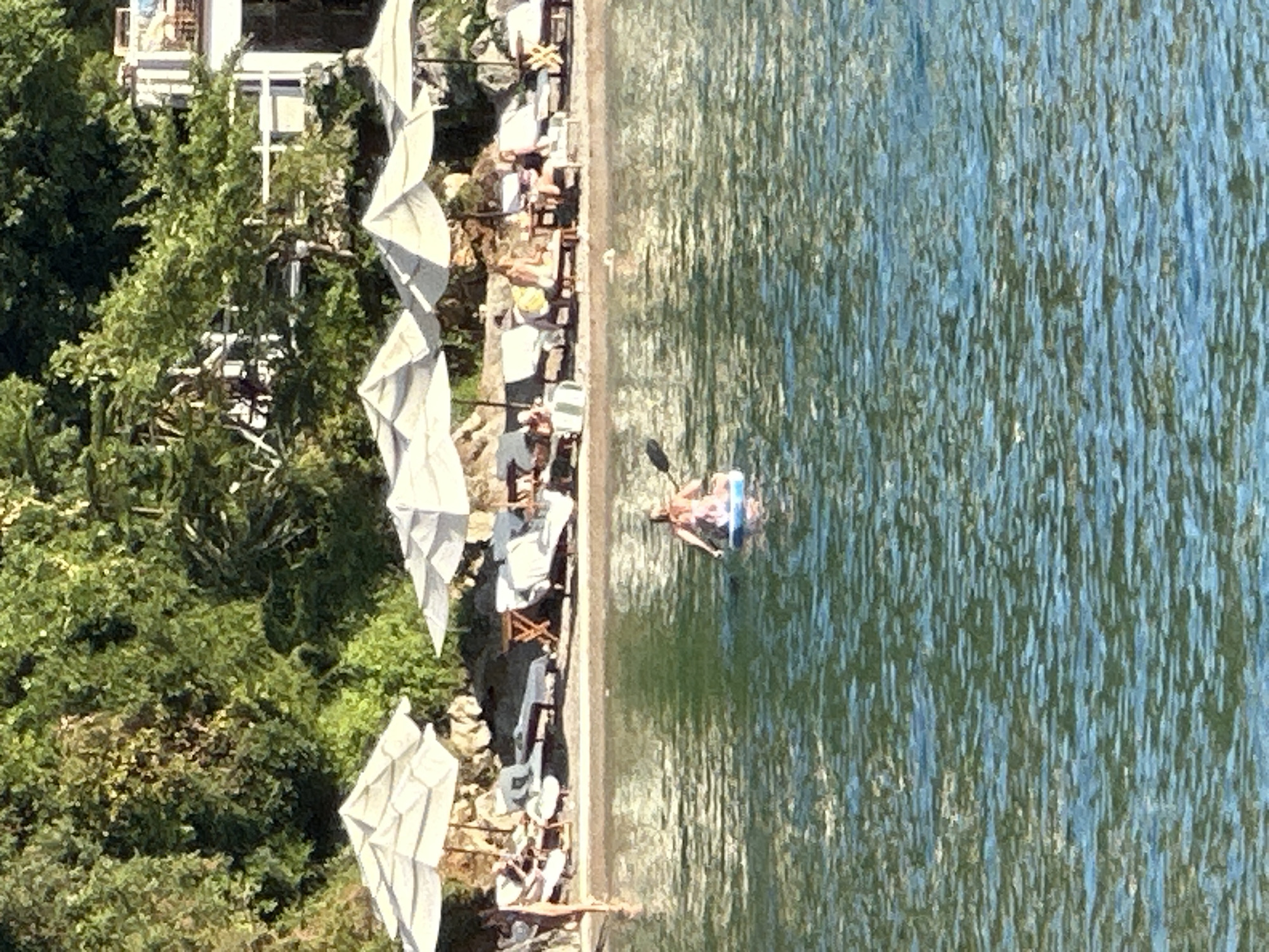 The image captures a tranquil scene at a beachside resort in Budva, Montenegro. In the foreground, the shimmering blue water of the Adriatic Sea is visible, with ripples and reflections indicating movement. A person is paddleboarding on a blue board, holding a paddle, their back to the camera. The middle ground features a sandy beach lined with white umbrellas and lounge chairs, some of which are occupied by people. The beach is backed by lush green trees and vegetation, with a white building structure visible in the upper right corner, suggesting accommodation or a restaurant. The overall atmosphere is one of relaxation and leisure, characteristic of a Mediterranean vacation spot.