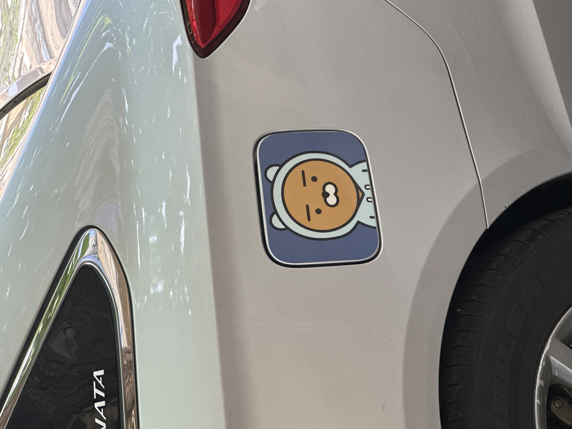 A close-up photograph captures the rear-right side of a silver or light grey sedan, specifically focusing on the fuel door, in the city of Unknown, Unknown. The fuel door is covered by a rectangular panel with rounded corners, featuring a cartoon character resembling Ryan from Kakao Friends. This character has an orange-brown face with small black dot eyes and a white, open-mouthed snout, and is depicted wearing a light blue hooded sweatshirt against a muted blue background. To the left, a partial view of a black rear window trim with a chrome outline is visible, displaying the white letters "NATA," indicative of a "SONATA" emblem. The upper portion of a red taillight lens is visible on the far right of the frame. In the lower left, the upper section of a black car tire and a silver multi-spoke wheel rim, showing signs of wear, are present. The car's paint surface exhibits reflections consistent with an outdoor, sunlit environment, showing green foliage and sky. No individuals or specific actions are depicted.