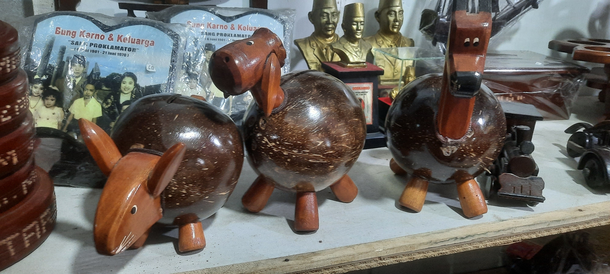 The image depicts a close-up of several wooden handicraft items displayed on a shelf. In the foreground, there are three animal-shaped objects made from coconuts and wood. The leftmost object appears to be a donkey with wooden ears and legs attached to a coconut body. To its right, a horse-shaped figure is made from a coconut body with wooden legs and a wooden head and neck. The third animal, also a horse, is situated on the right side of the frame, with a similar construction of coconut body and wooden extremities.
In the background, there are framed pictures and busts. One framed picture on the left shows a family portrait, with text that appears to read "Bung Karno & Keluarga" and "SAM PROKLAMATOR". Behind the animal figures, several golden busts of men in traditional hats are visible, possibly historical figures. A small wooden train is positioned to the right of the horse figures, and further to the right, a partial view of a wooden car can be seen. The overall setting suggests a handicraft shop or market stall. The image does not provide any specific visual cues that would allow for the precise location within Blitar, Indonesia, to be determined.
