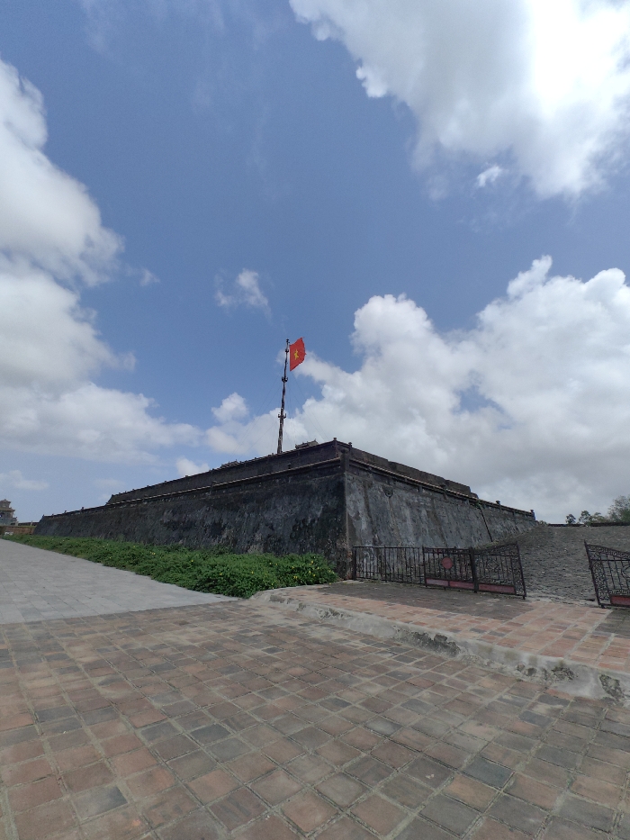 An outdoor, eye-level wide shot depicts a large, weathered stone fortress wall, or rampart, under a partly cloudy blue sky in Phú Xuân Ward, Vietnam. The wall, dark gray and appearing constructed of blocks, extends horizontally across the middle of the frame from left to right, turning at a corner on the right. A prominent multi-segmented flagpole is mounted atop the wall to the left of the corner, flying a red Vietnamese flag with a central yellow five-pointed star.
In the foreground, the ground is paved with reddish-brown square tiles. A raised, light-colored curb separates this area from a darker, possibly gray, paved path that runs diagonally towards the upper left, parallel to the wall. A strip of green ground cover, possibly low shrubs or vines with scattered yellow flowers, borders the base of the fortress wall on the left side of the frame. To the right, a dark metal fence with decorative scrollwork separates the tiled foreground from an upward-sloping, dark-colored stone or paved ramp leading to the top of the fortress. No people, discernible actions, or interactions are visible in the scene. The overall lighting suggests a bright, sunny day.