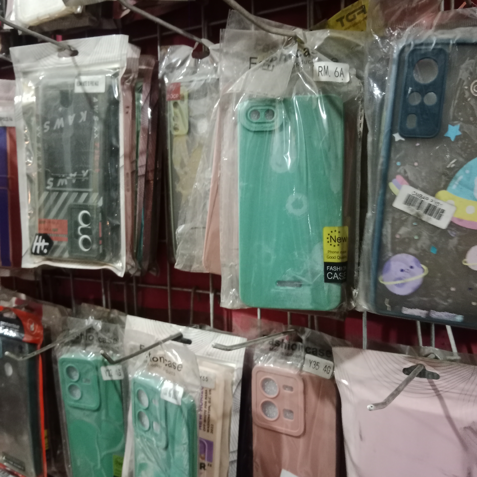 The image shows a close-up, eye-level, horizontal shot of a retail display featuring numerous mobile phone cases. The cases are individually packaged in clear plastic and are hung on a metal rack. Various colors and designs are visible, including solid green, pink, and patterned cases with illustrations. Some packaging includes labels with prices such as "RM. 6A" and model numbers like "Y35 4G." A label with "KAWS" branding is also visible on one of the cases. The overall environment suggests a small shop or market stall. There are no people or animals present in the image.