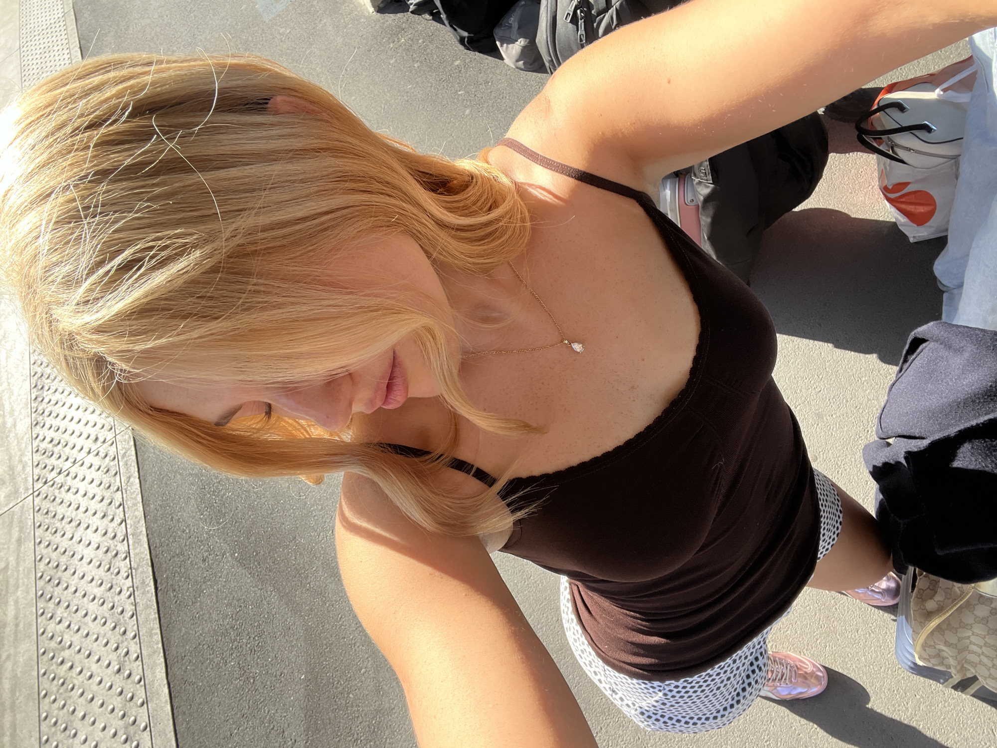 A young woman stands outdoors on a textured pavement surface, possibly in Paris, France. She is captured from an overhead angle in a selfie, showcasing her blonde hair, fair skin, and a brown tank top with thin straps. A delicate gold necklace adorns her neck. She is wearing a white patterned skirt and metallic pink sneakers. Adjacent to her are other individuals and objects including a black backpack, a white bag with a red graphic, a light blue denim garment, and a handbag with a monogram design. The setting suggests a public space, such as a sidewalk or train platform, given the textured surface. Bright sunlight illuminates the scene, casting shadows and accentuating details.