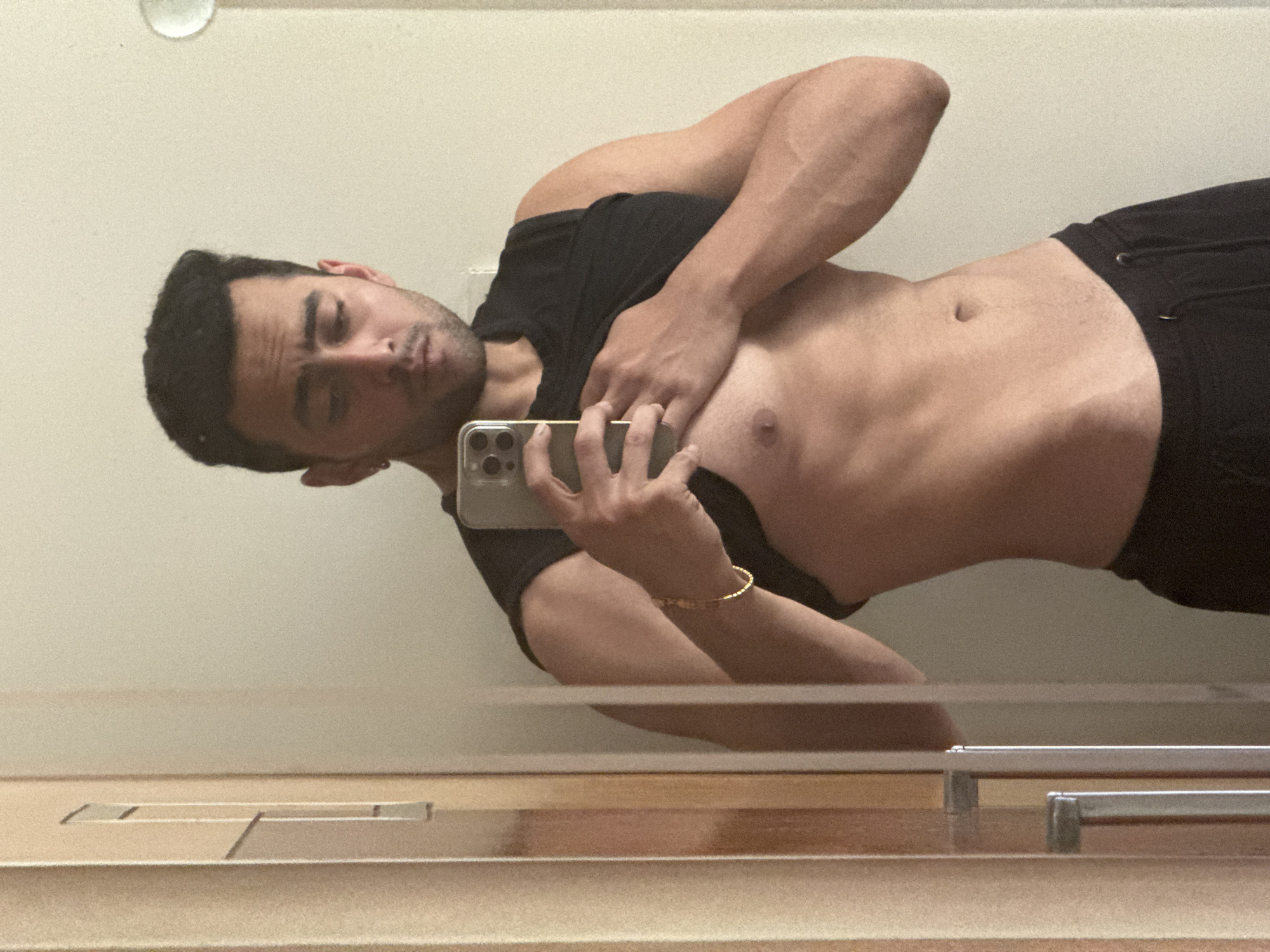 A person with dark hair, wearing a black t-shirt and black shorts, is taking a selfie in a bathroom mirror. The person's torso is exposed, and they are holding a smartphone in their right hand. A gold bracelet is visible on their wrist. The background is a plain, light-colored wall with a medicine cabinet below the mirror. The location cannot be determined from the image, so Kolkata, India cannot be verified.