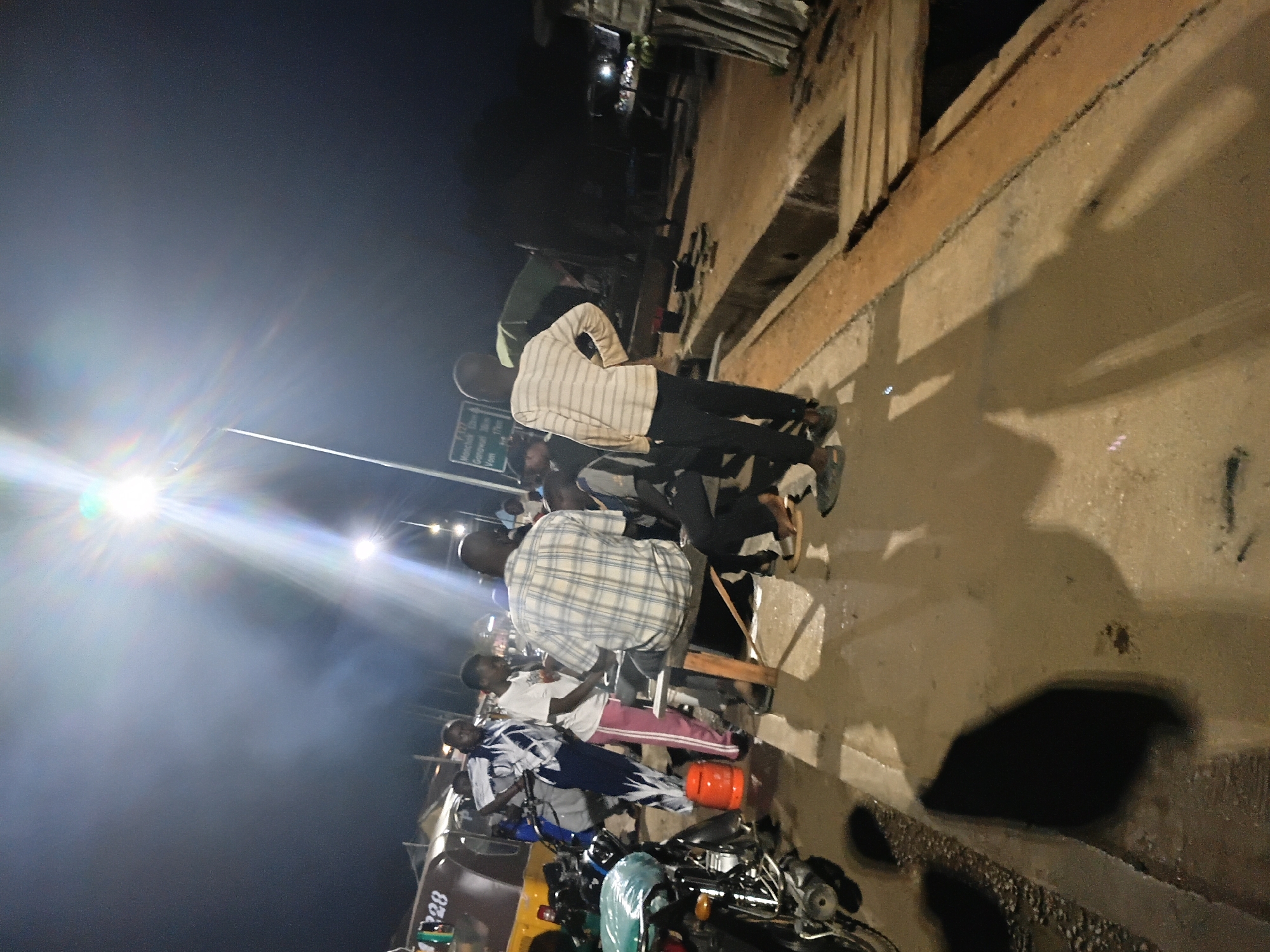 This image, taken at night in Bukuru, Nigeria, depicts a group of people gathered around a brightly lit street lamp. The scene appears to be on a roadway or a public gathering area. In the foreground, a motorcycle is parked, its front tire and fender visible. To the left, a yellow and brown three-wheeled vehicle, possibly a tuk-tuk or commercial tricycle, is partially visible with the number "28" on its side.
Several individuals are present. A man in a plaid shirt is seated on a wooden bench. Another person stands behind him, leaning forward. A person in a white and blue patterned garment stands near the three-wheeled vehicle, and another individual in a white t-shirt and pink pants is also present. Further back, a person wearing a striped long-sleeved shirt and dark pants stands facing away from the camera, with hands on their hips. Shadows are cast on the ground by the individuals and objects, indicating a strong light source from above.
The environment suggests an urban or semi-urban setting at night. The ground is wet or damp, reflecting the light. In the background, other vehicles and structures are dimly visible, along with streetlights and possibly distant buildings. A signpost with text is discernible in the background, but the details are illegible. The overall impression is a snapshot of a nocturnal social gathering or waiting area in Nigeria.