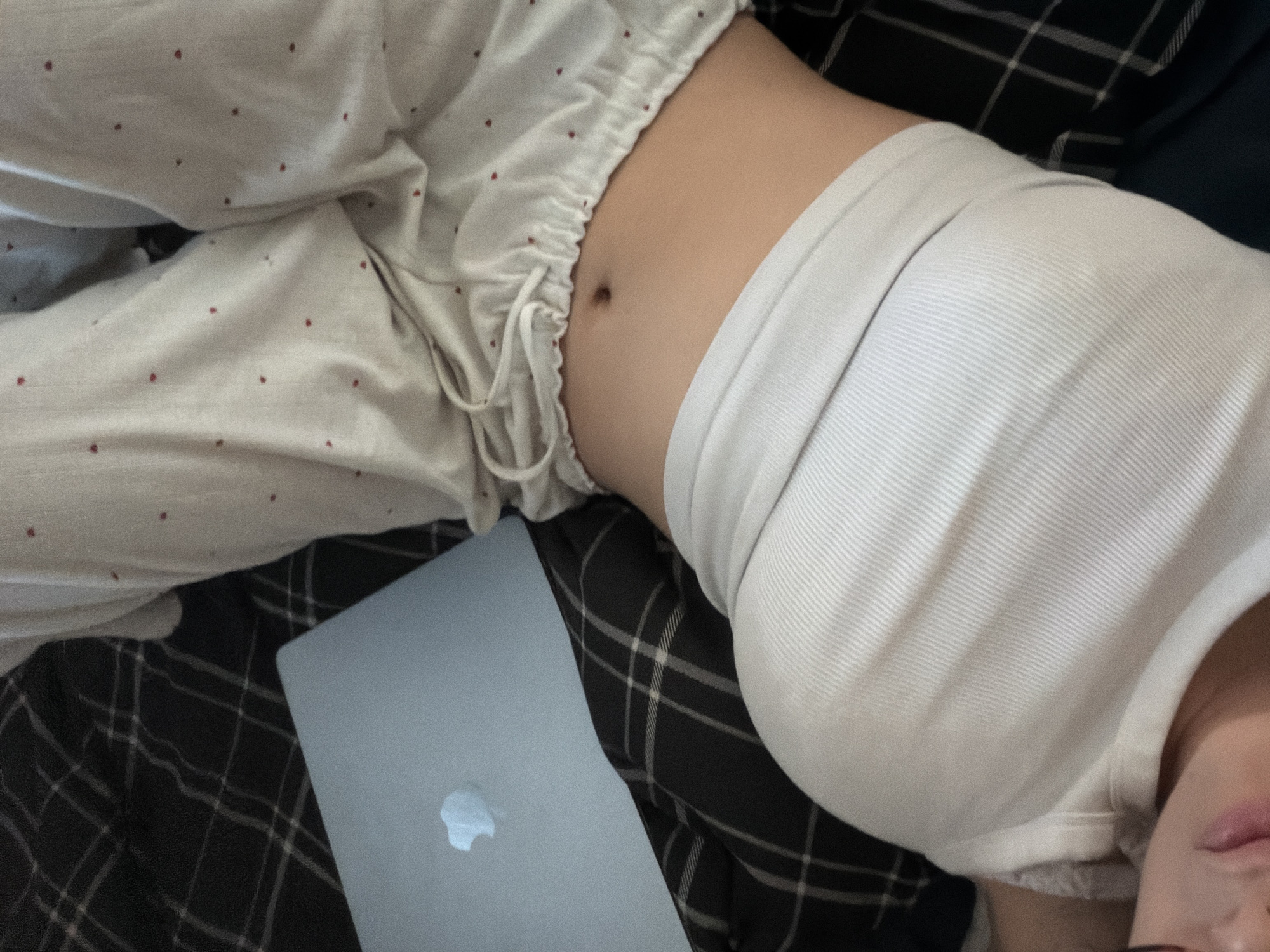 The image shows a close-up, low-angle view of a person lying down on a bed with a laptop nearby. The person is wearing a white ribbed tank top and light-colored pajama pants with small red dots. Their stomach is visible, and their face is partially cropped at the bottom left of the frame. The bed is covered with a dark plaid blanket and the person's pajama pants. The laptop, a silver MacBook, is open and placed on the blanket to the left of the person. There is no indication of the location being Milan, Italy.