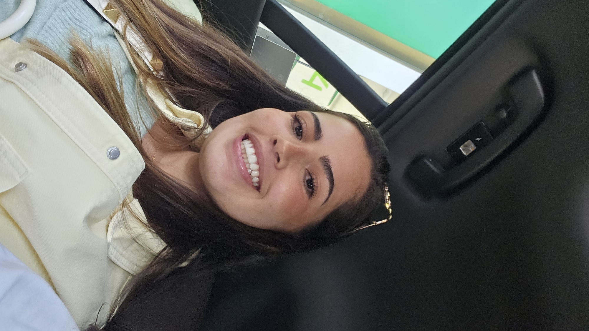 A young woman with dark hair and light skin is smiling at the camera. She is wearing a light yellow jacket over a light blue top. She is inside a vehicle, with a black interior and a sunroof. A handle with control buttons is visible above her. The location is Mexico City, Mexico. There is a green sign in the background.