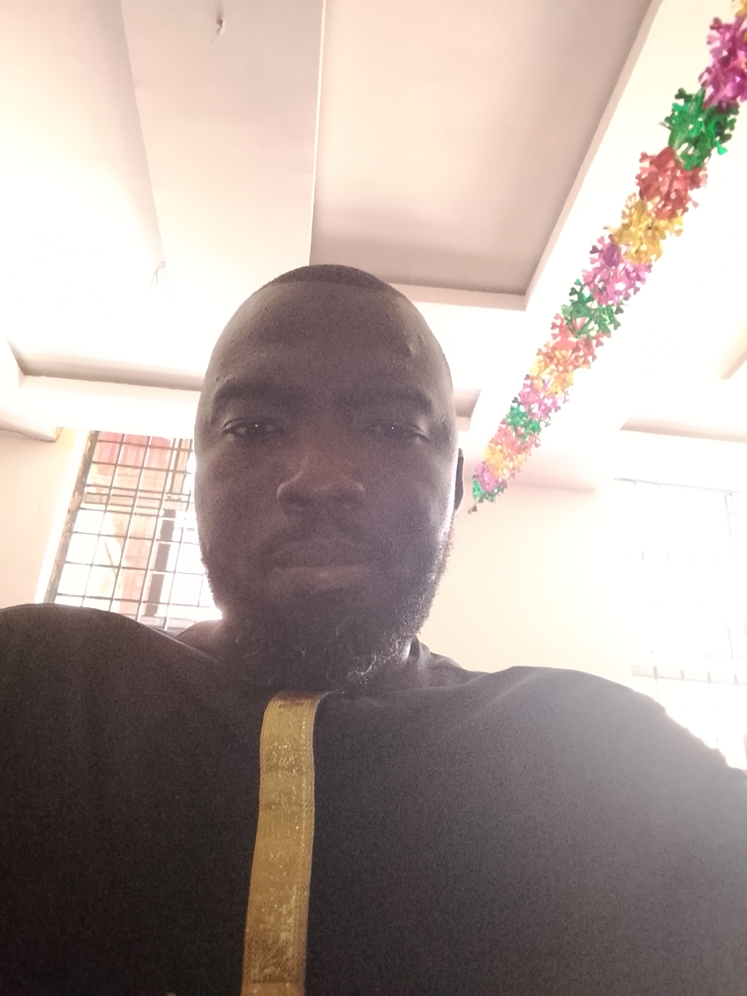 A close-up selfie of a Black man with a shaved head and a beard. He is wearing a black shirt with a gold stripe on the shoulder. The background shows a white ceiling with recessed lighting and a colorful garland hanging from it. A window with a metal security grill is visible on the left. The photo was likely taken indoors in Jos, Nigeria.