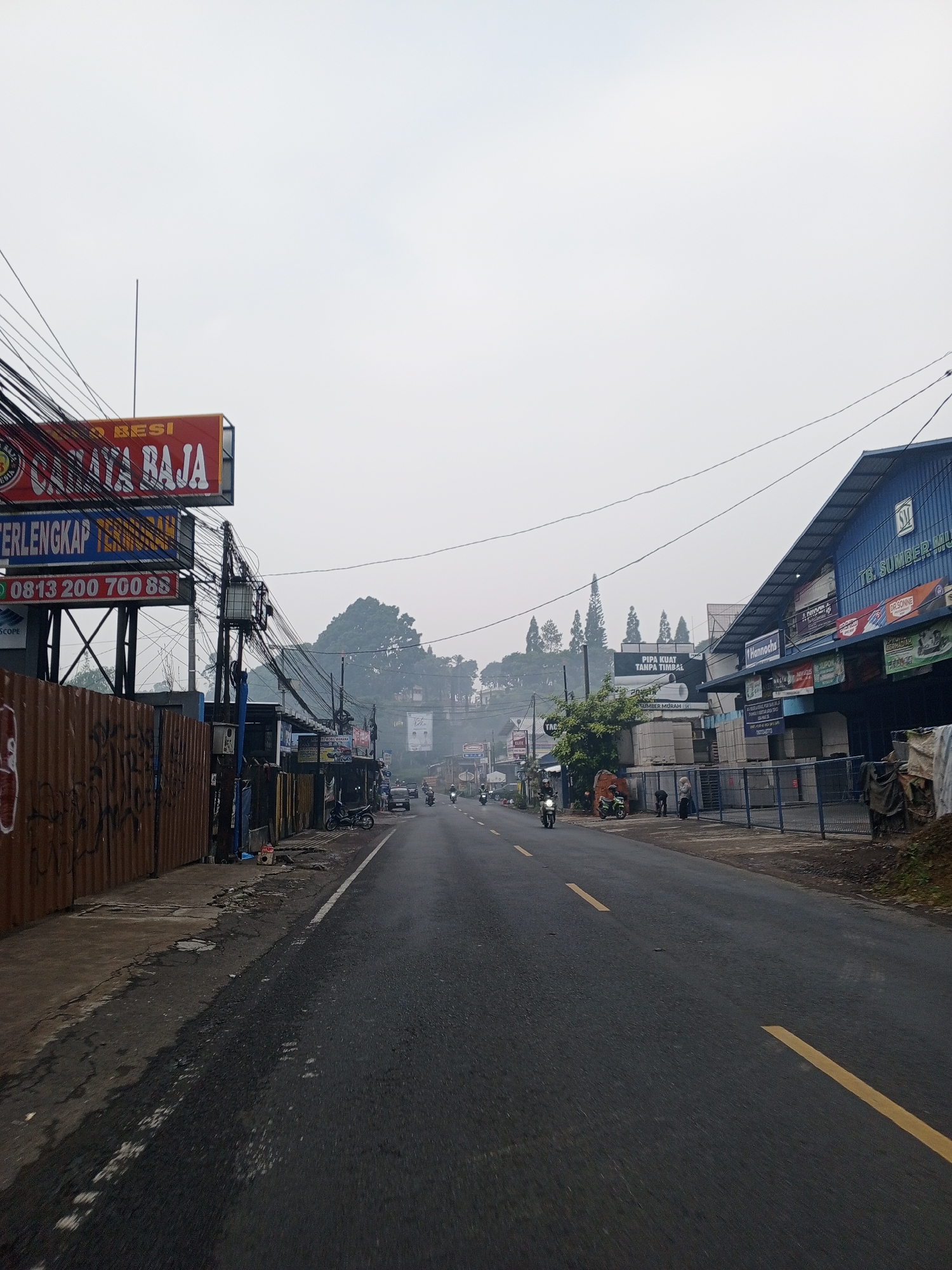 The image depicts a roadside scene in Dambulla, Sri Lanka, characterized by a slightly hazy, overcast sky. A paved road with a yellow dividing line runs through the center of the frame, with a few motorcycles and a car visible in the distance. On the left side of the road, a corrugated metal fence with graffiti is present, alongside a tall utility pole laden with a complex network of wires and electrical boxes. Above the fence, a red and yellow sign with text in Indonesian reads "MAKMUR JAYA BESI TERLENGKAP TERMURAH" and a phone number "0813 200 700 88". Adjacent to this, a blue sign is visible. Further down the road on the left, a building with several windows and a sign that reads "DS KOP" is present.
On the right side of the road, a long, blue-roofed building dominates the view. This building has multiple signs, including one that says "TB SUMBER MURAH". Other signs on this building include "Hannochs", "DR.SONNE", and other unreadable text. In front of this building, a blue metal fence encloses an area with some stacked building materials. Several people are visible near the fence, some walking and others standing. A green motorcycle is parked on the side of the road near these individuals. Further down the street on the right, more buildings and signs are visible. The environment suggests a commercial or industrial area on the outskirts of a city, with dense vegetation on a hillside in the background. The overall impression is one of a moderately busy street in a tropical or subtropical climate.