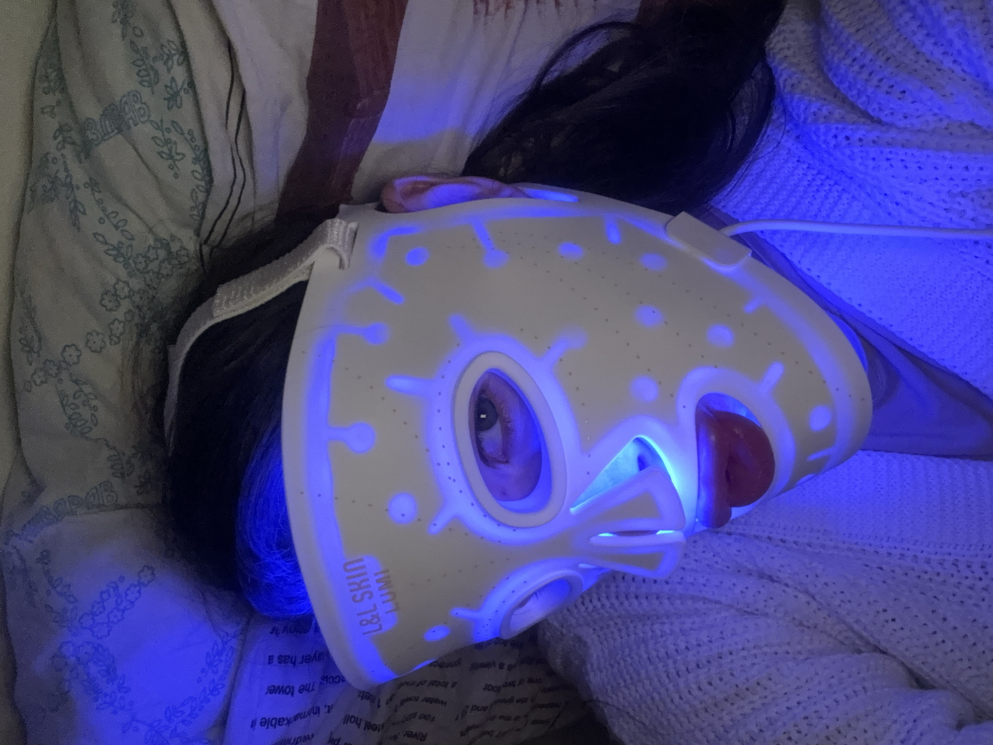 A woman lies on a bed with a blue LED light therapy mask on her face. She is on top of a white knit blanket. Her head is resting on a pillow with a floral pattern and some writing is visible on the pillow. The mask has the brand "L&L SKIN LUMI" printed on the side.